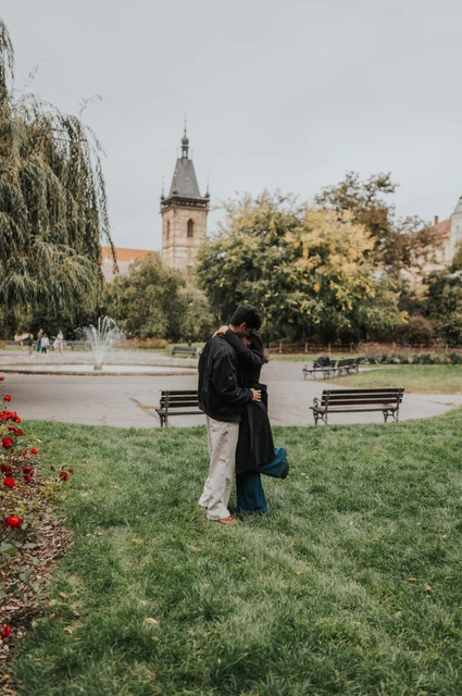 Prague couple photoshoot: an embrace beside the flowerbeds Couple embracing on the grass beside a flowerbed with the park fountain behind them during their Prague couple photoshoot.