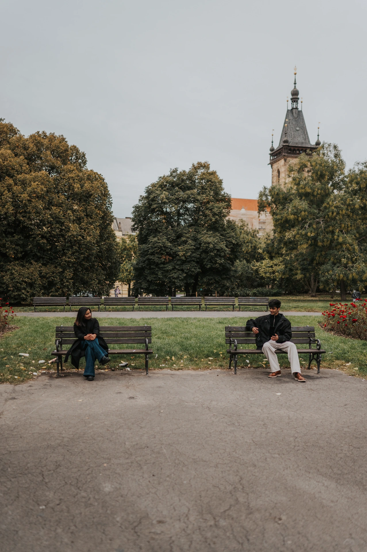 Couple sitting apart on two park benches with green lawns and the tower above them during their Prague couple photoshoot.