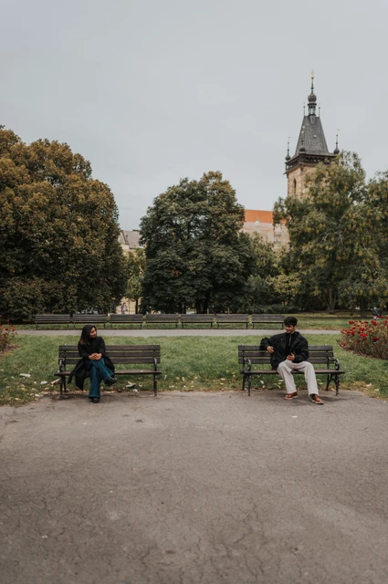 Prague couple photoshoot: two benches framed by the Charles Square trees Couple sitting apart on two park benches with green lawns and the tower above them during their Prague couple photoshoot.