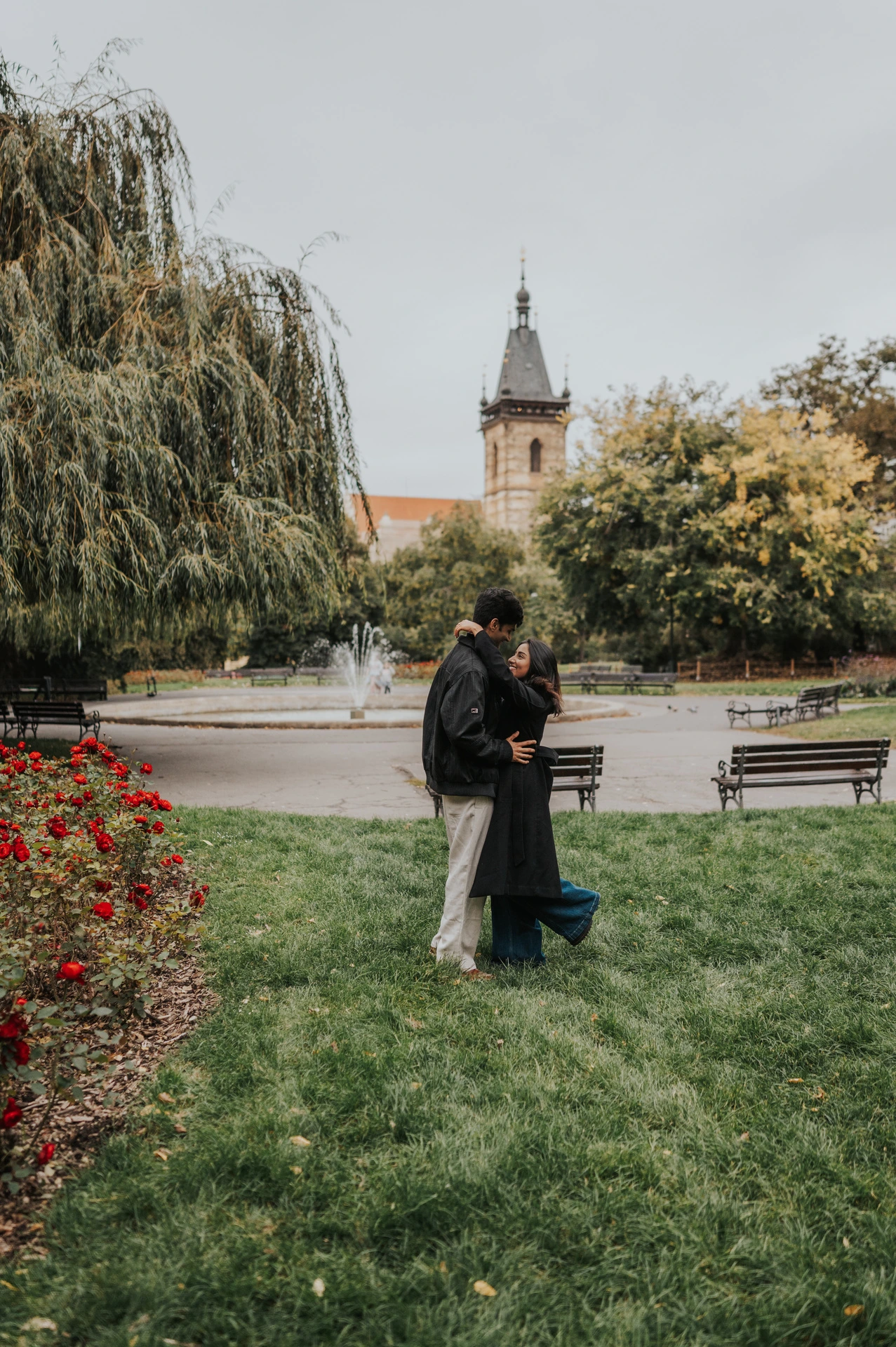 Couple standing close together on the lawn beside flowers and park benches during their Prague couple photoshoot.