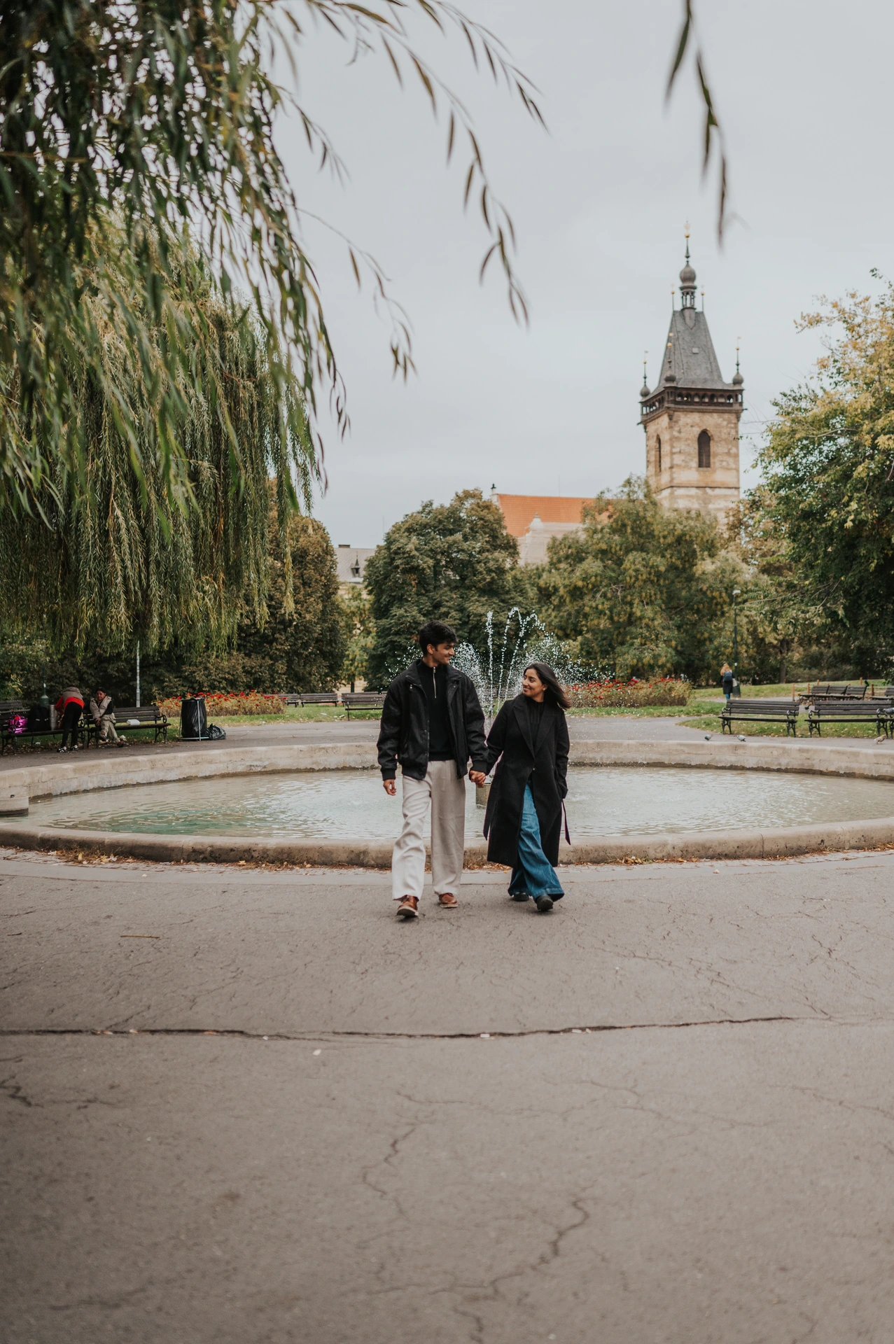 Couple walking together beside the fountain and flowerbeds in Charles Square Park during their Prague couple photoshoot.