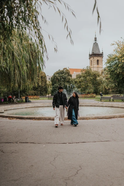 Prague couple photoshoot: walking past the Charles Square fountain Couple walking together beside the fountain and flowerbeds in Charles Square Park during their Prague couple photoshoot.