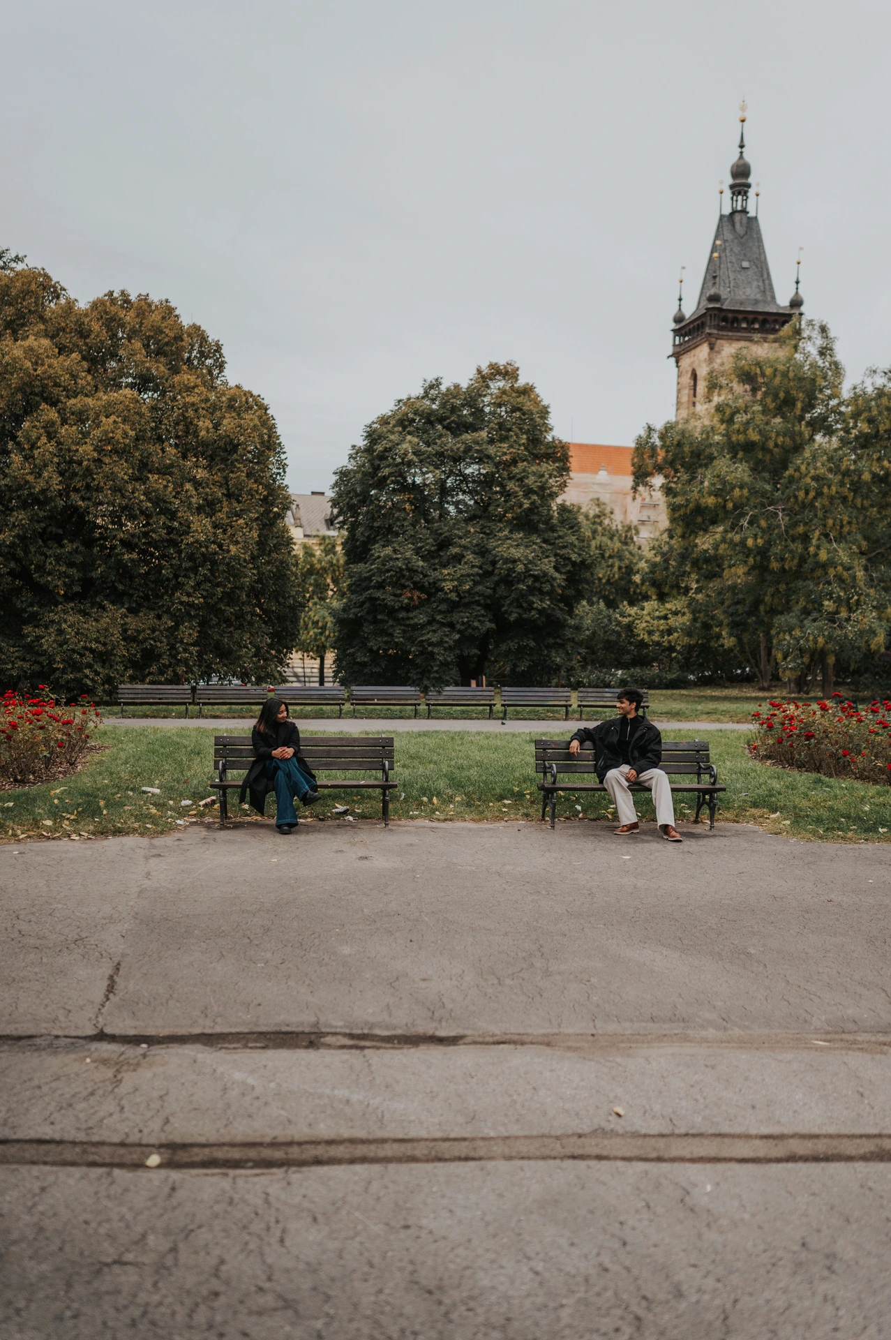 Wide view of the couple sitting on separate park benches with trees and the New Town Hall tower behind them during their Prague couple photoshoot.