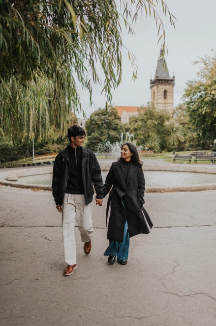 Couple walking hand in hand along a tree-lined path in Charles Square Park with the New Town Hall tower behind them during their Prague couple photoshoot.