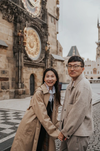 Couple smiling at the camera and holding hands with the Astronomical Clock behind them during their Prague couple photoshoot.