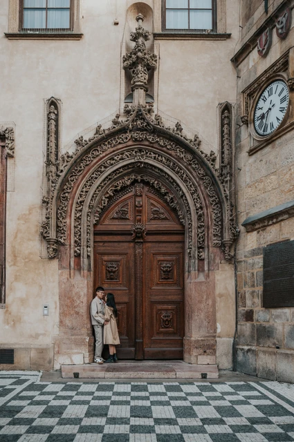 Full-length portrait of the couple standing close in front of the Old Town Hall's tall wooden door during their Prague couple photoshoot.