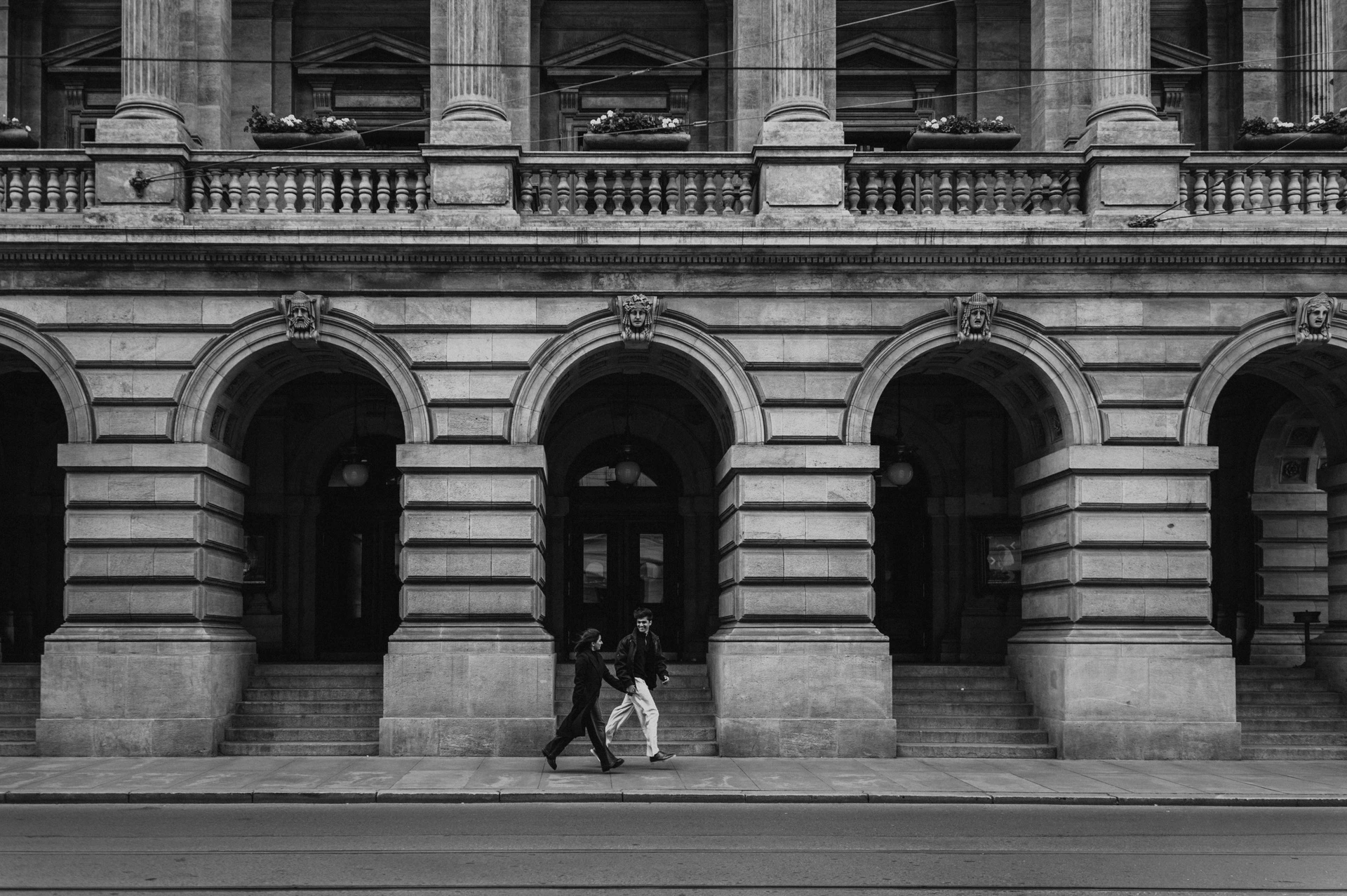 Black-and-white wide photograph of the couple walking in front of the National Theatre arcade during their Prague couple photoshoot.