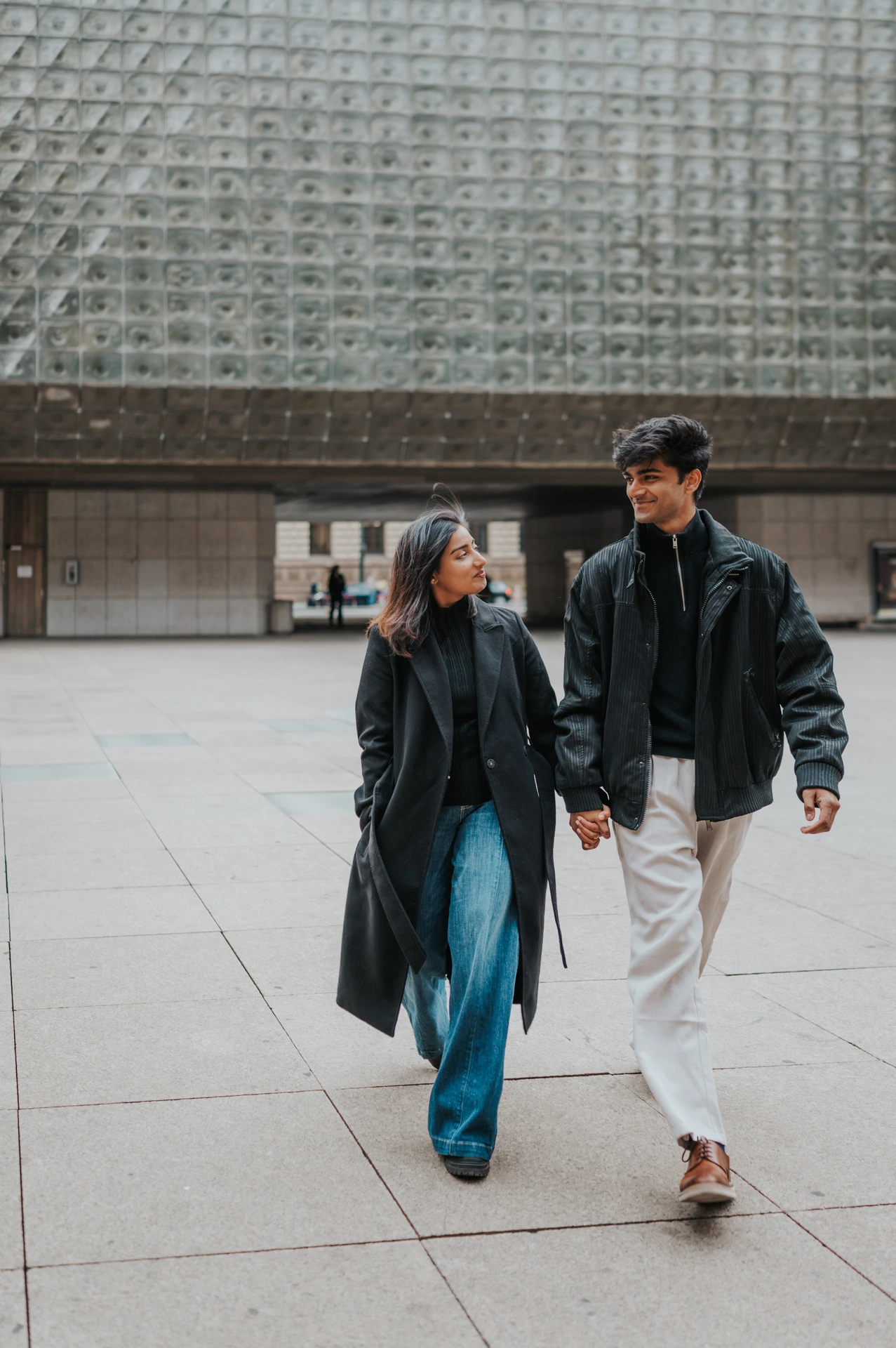 Couple walking hand in hand in jeans beside a textured modern facade during their Prague couple photoshoot.