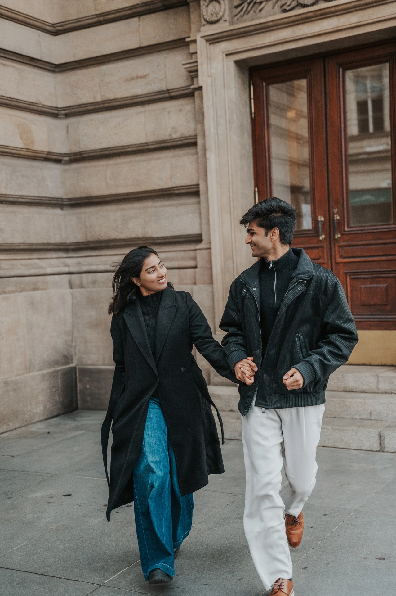 Couple walking hand in hand beside the National Theatre's stone wall and wooden doors during their Prague couple photoshoot.
