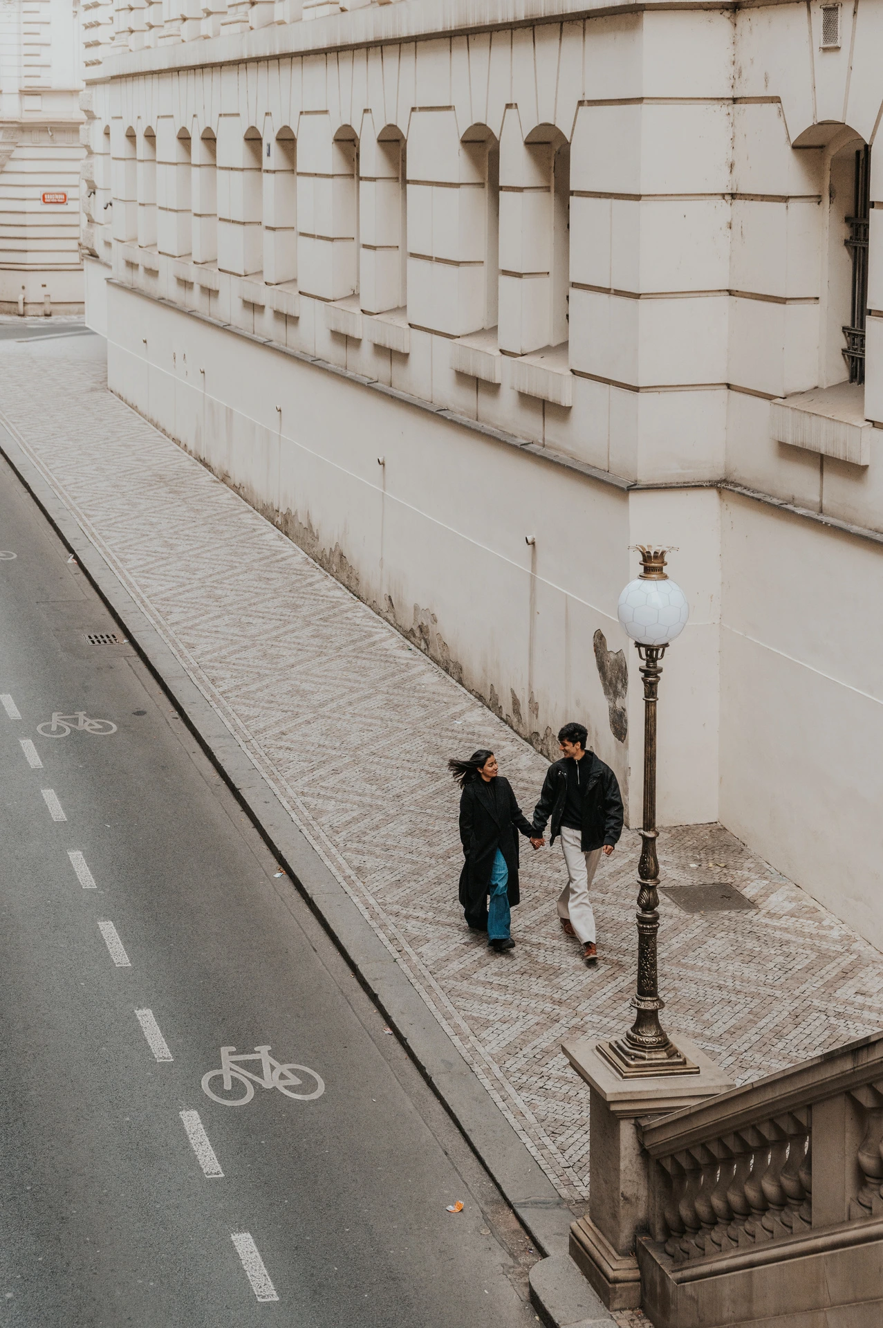 Couple holding hands on the pavement below a street lamp beside the theatre wall during their Prague couple photoshoot.