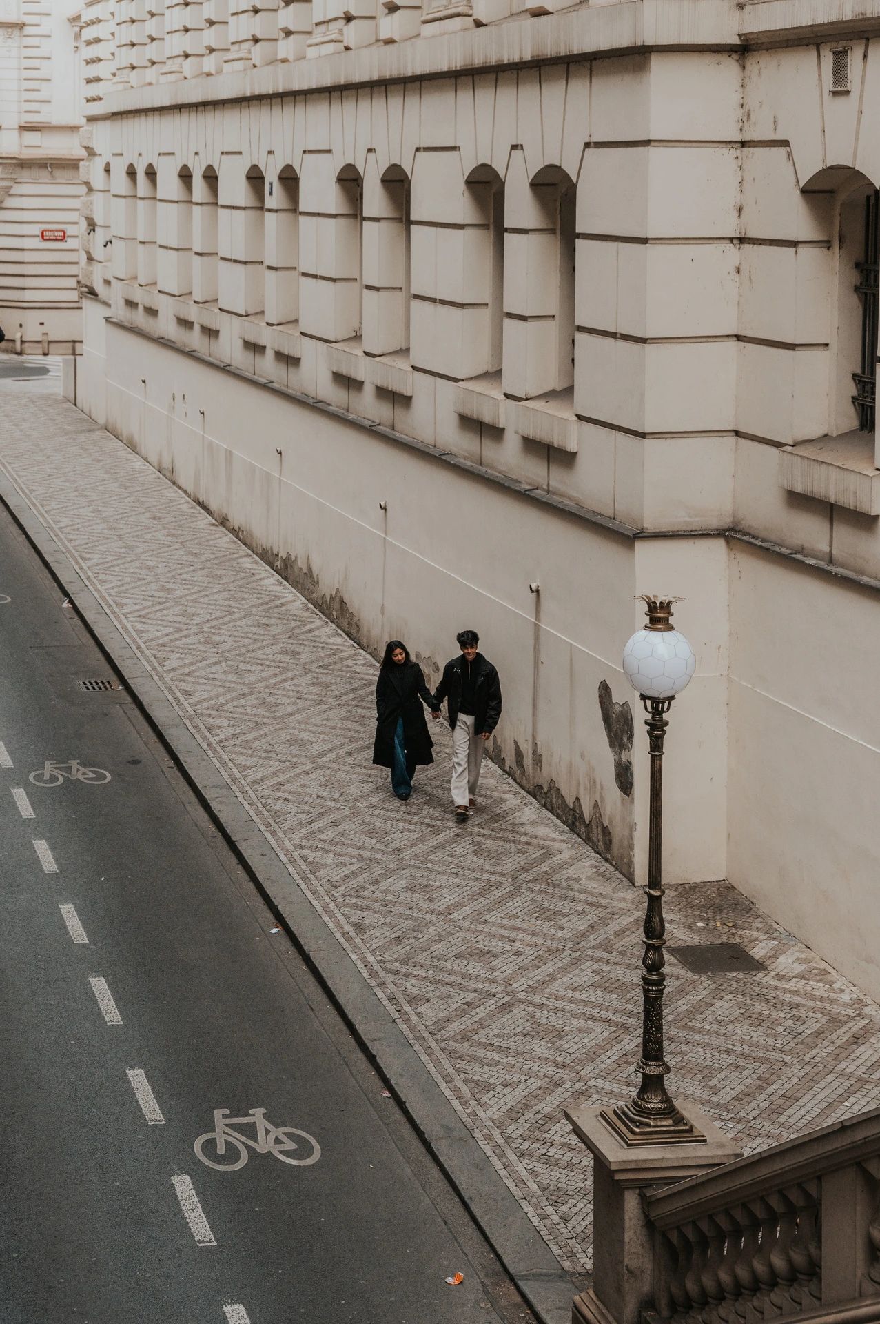 Overhead view of the couple walking beside the theatre's long stone wall and a street lamp during their Prague couple photoshoot.