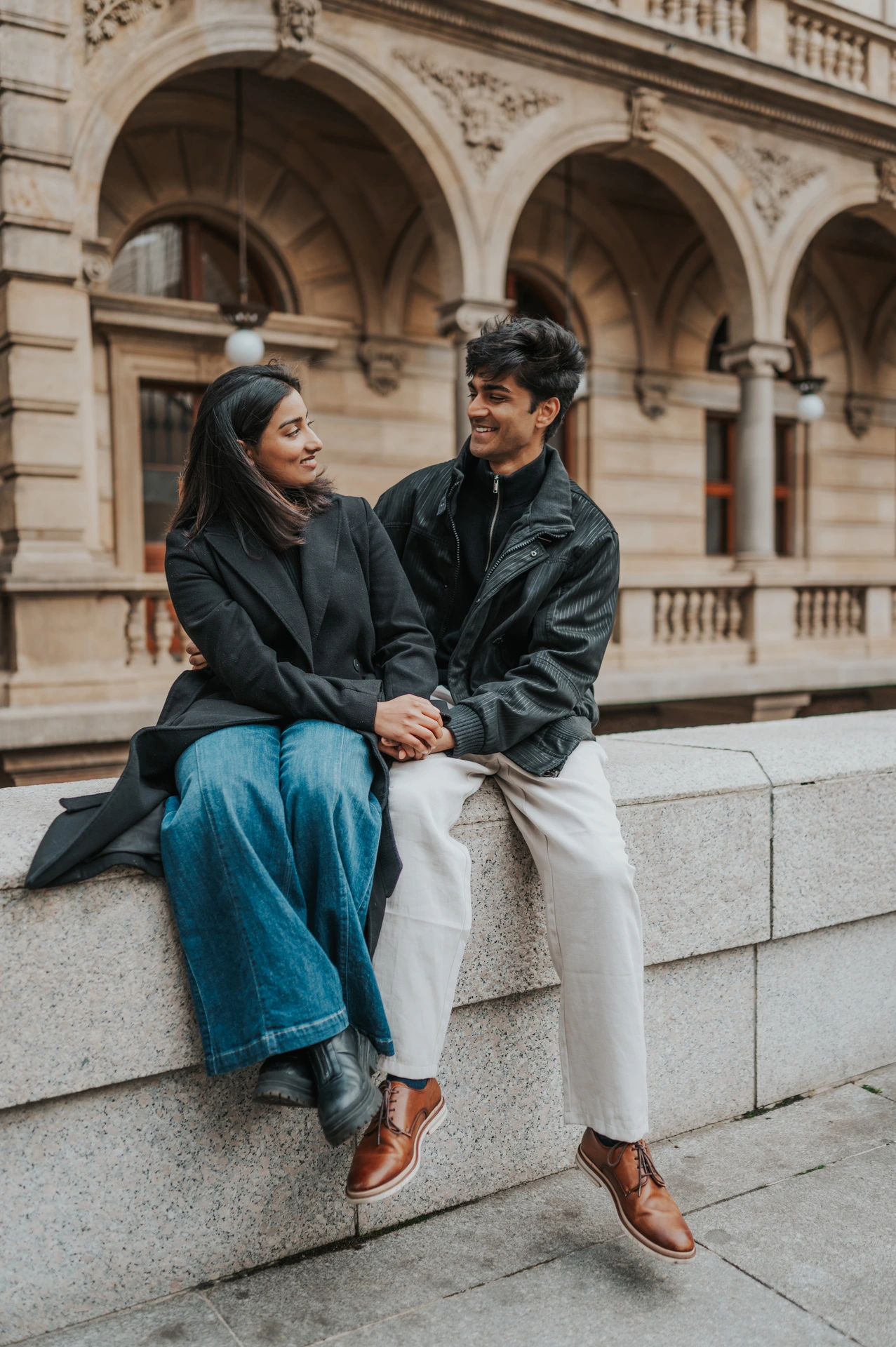 Couple smiling at each other while seated in front of the National Theatre's classical arches during their Prague couple photoshoot.
