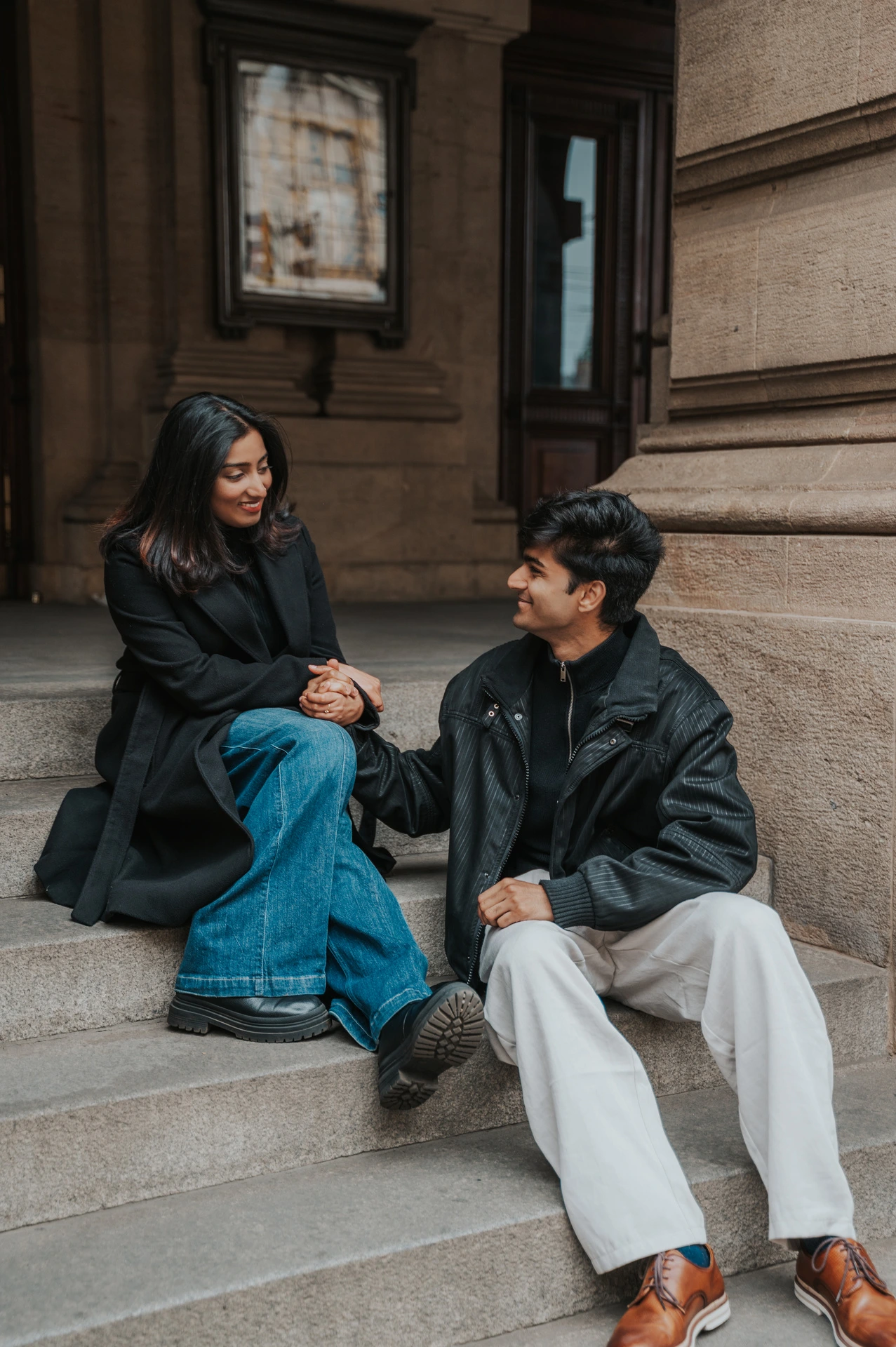 Couple holding hands and smiling at each other while sitting on the theatre steps during their Prague couple photoshoot.
