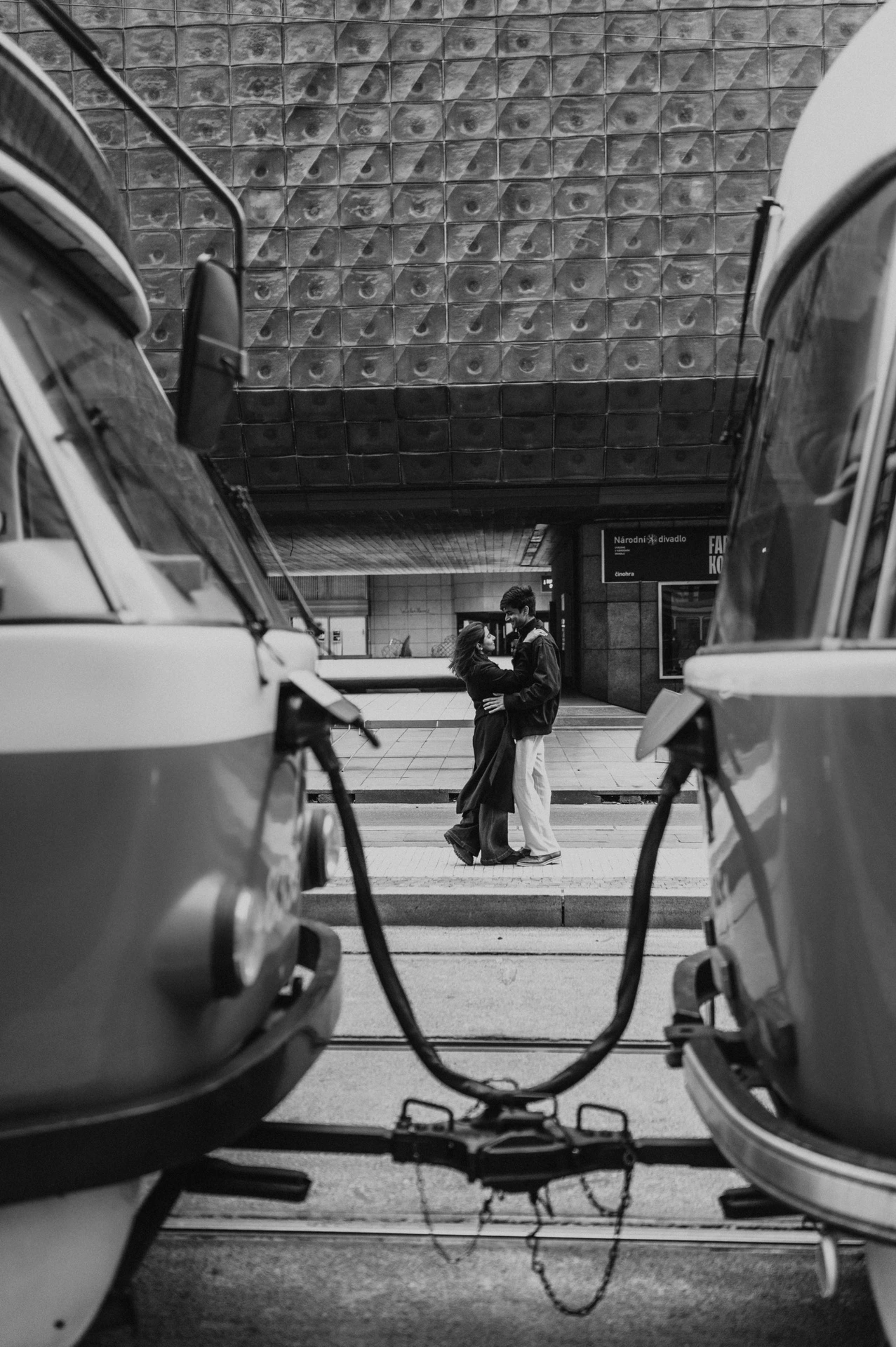 Black-and-white photograph of the couple embracing between two trams outside the theatre during their Prague couple photoshoot.