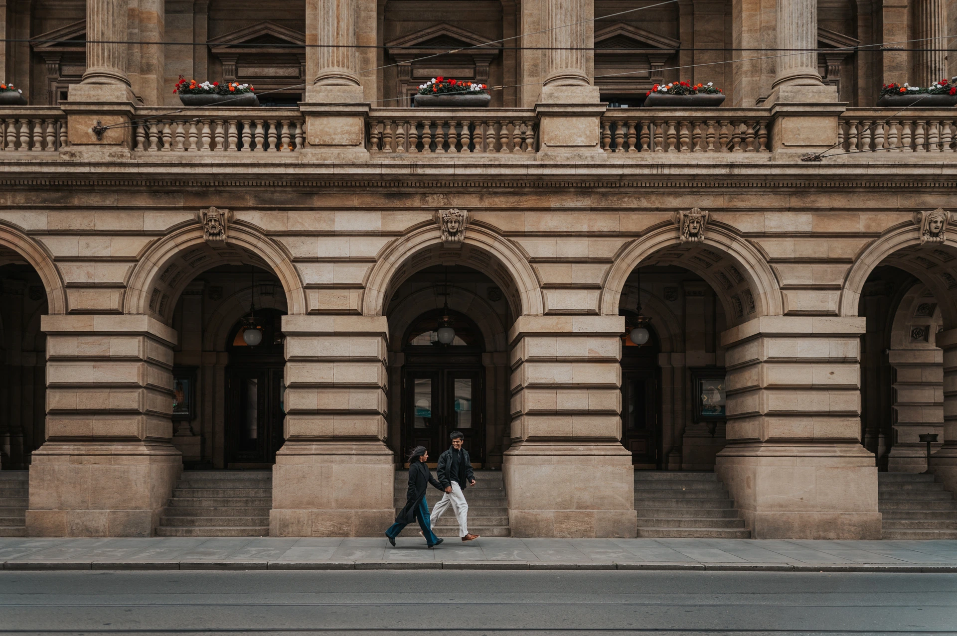 Couple holding hands beneath an arch in a wide view of the historic theatre facade during their Prague couple photoshoot.