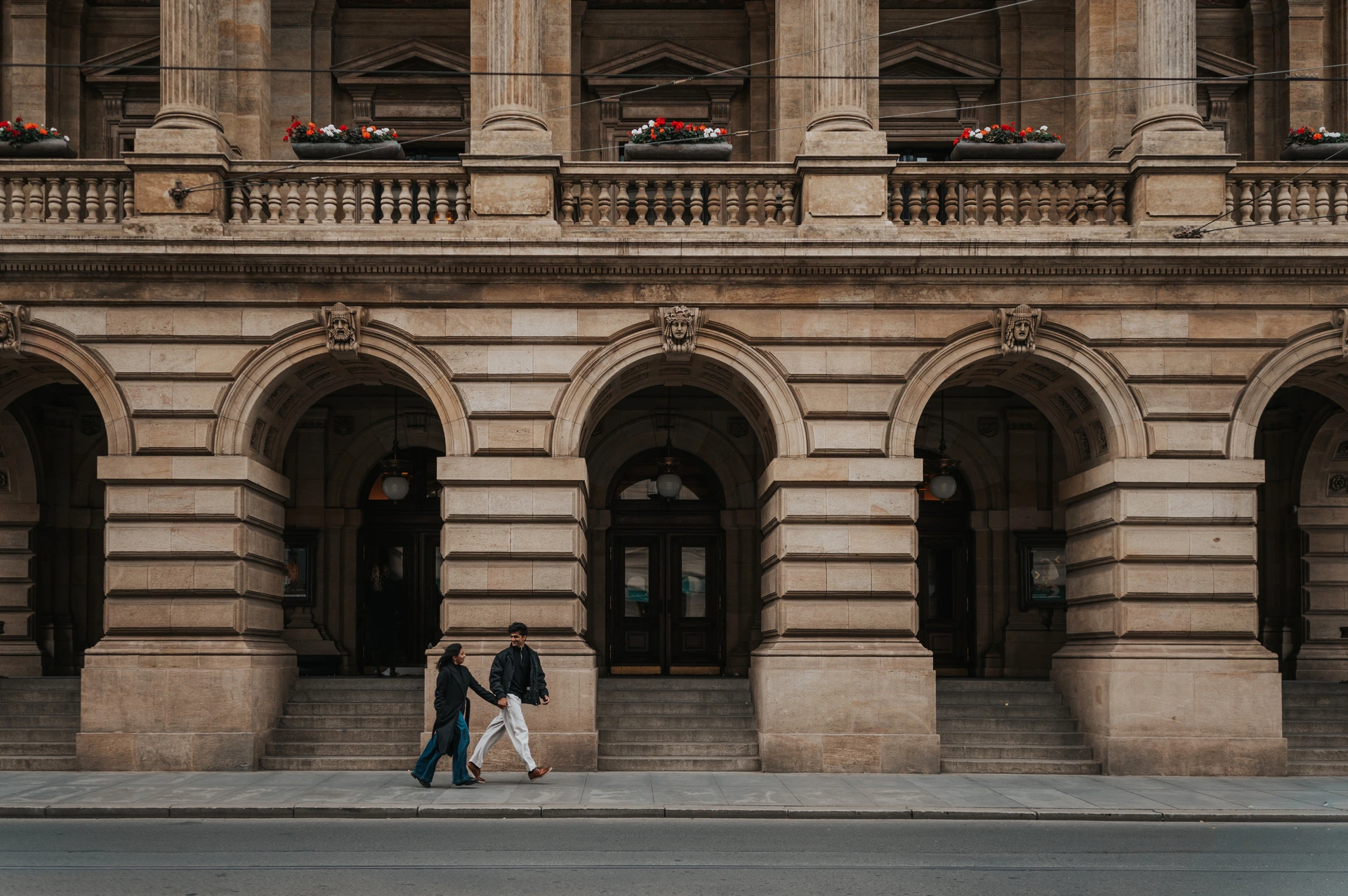 Prague couple photoshoot: walking beneath the National Theatre arches Wide view of the couple walking past the National Theatre's ornate arched facade during their Prague couple photoshoot.
