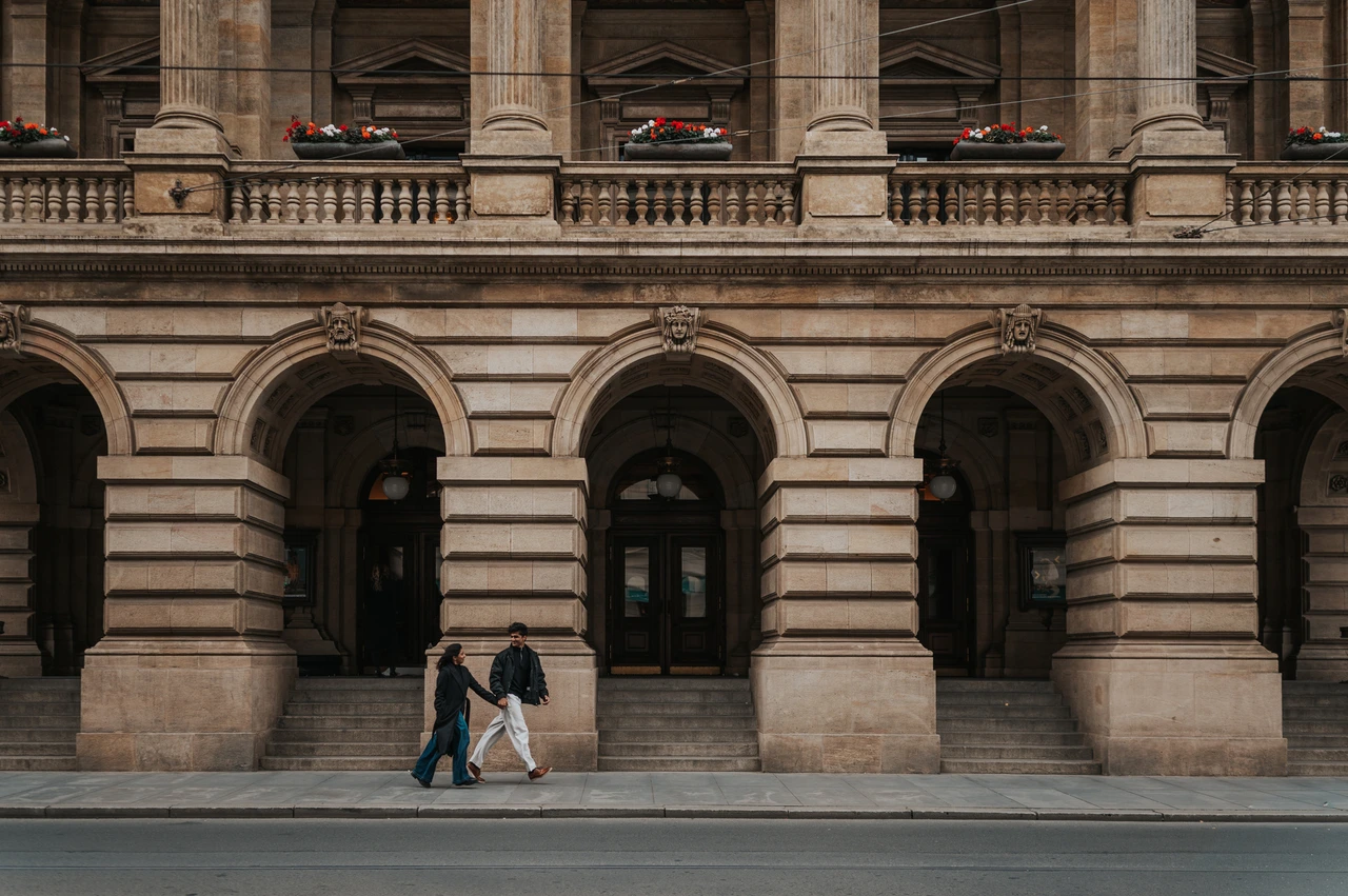 Prague couple photoshoot: walking beneath the National Theatre arches Wide view of the couple walking past the National Theatre's ornate arched facade during their Prague couple photoshoot.