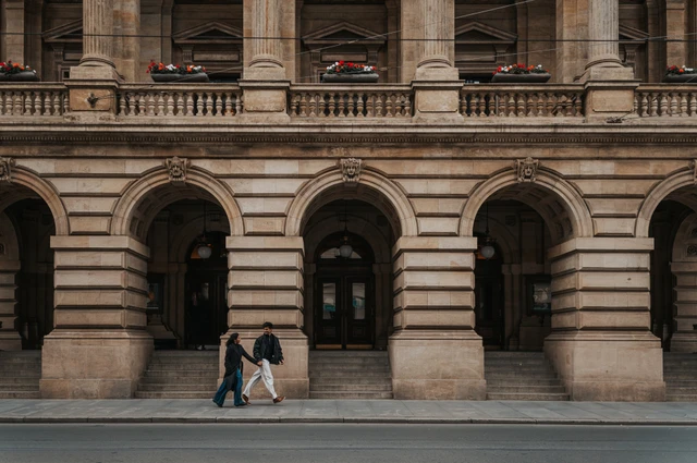 Wide view of the couple walking past the National Theatre's ornate arched facade during their Prague couple photoshoot.