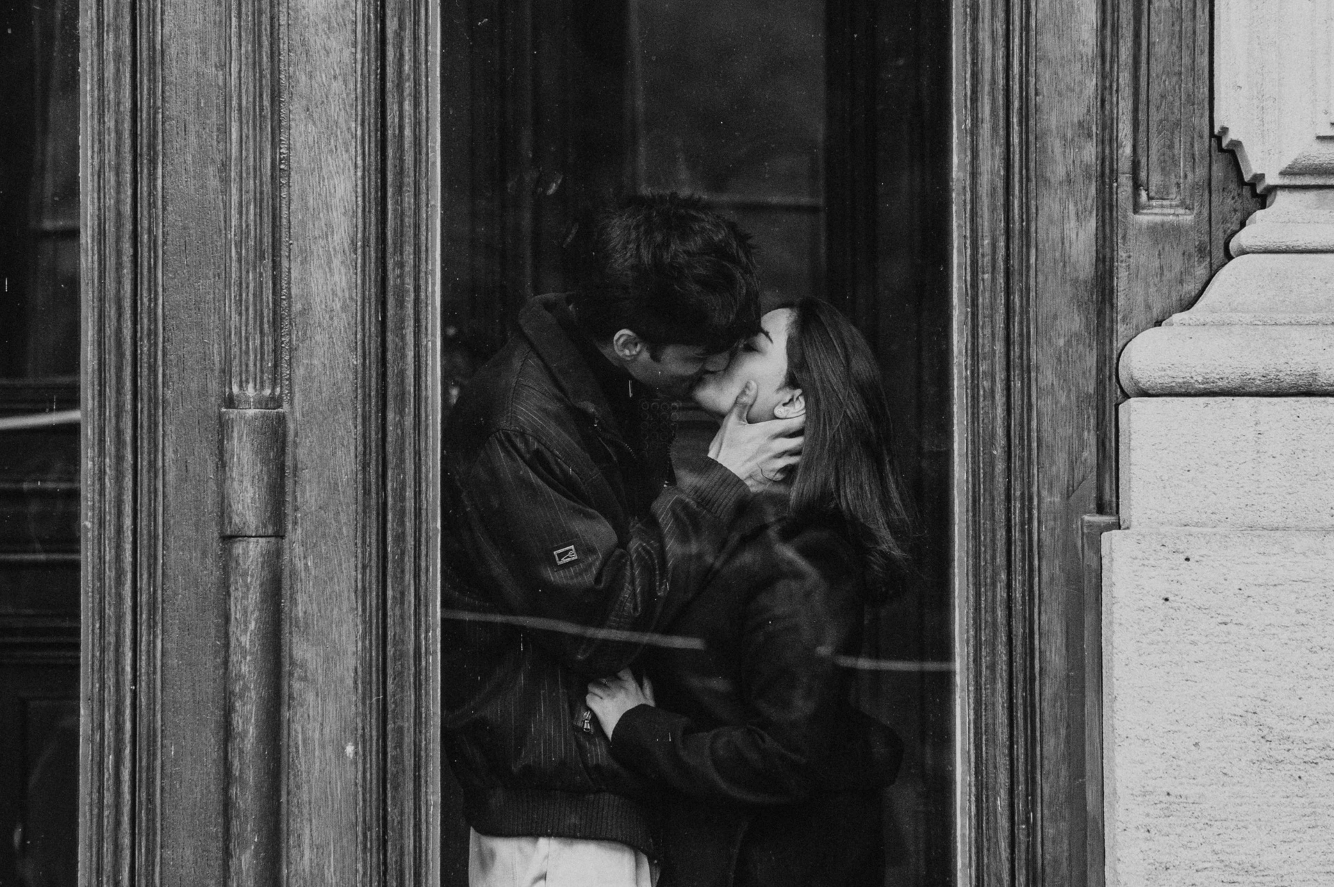 Black-and-white wide photograph of the couple kissing within an ornate wooden window frame during their Prague couple photoshoot.