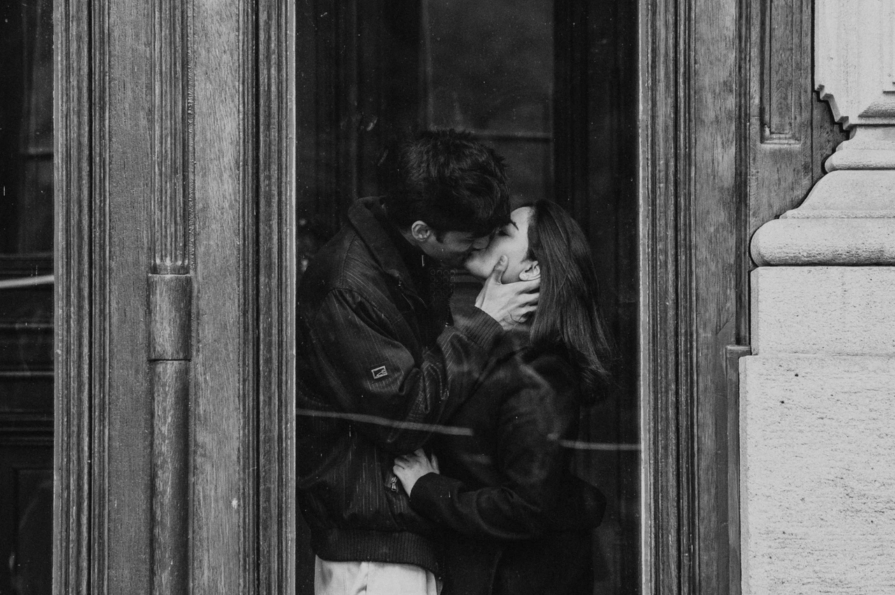 Prague couple photoshoot: a black-and-white window-frame kiss Black-and-white wide photograph of the couple kissing within an ornate wooden window frame during their Prague couple photoshoot.