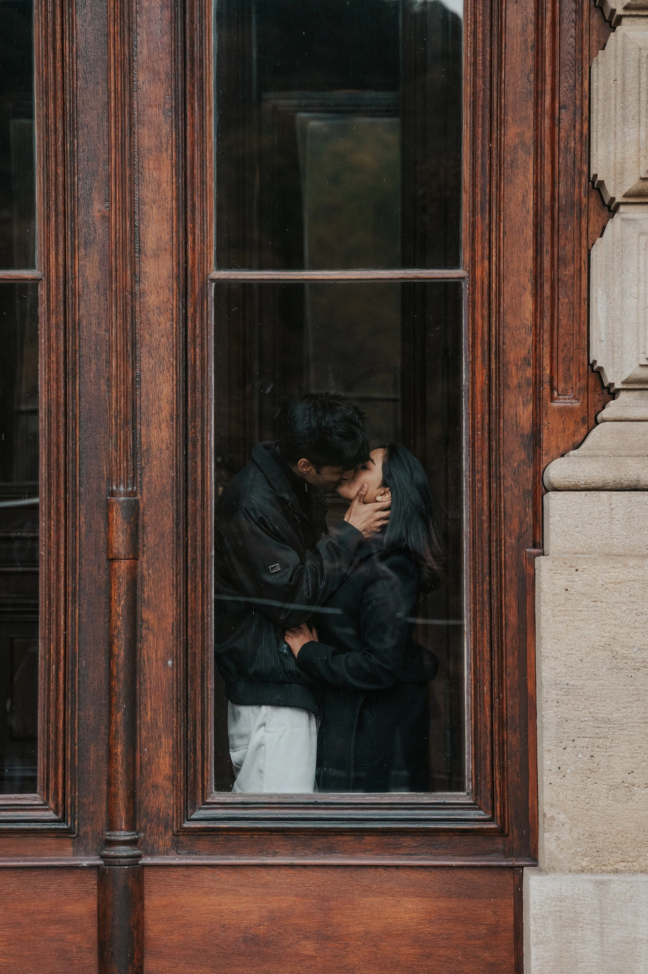 Couple embracing and sharing a kiss inside a tall wooden window frame during their Prague couple photoshoot.