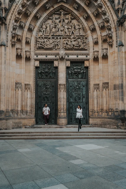 Prague photoshoot framed by the St. Vitus Cathedral entrance Distant full-length portrait of the couple beside the dark cathedral doors during their photoshoot at Prague Castle.
