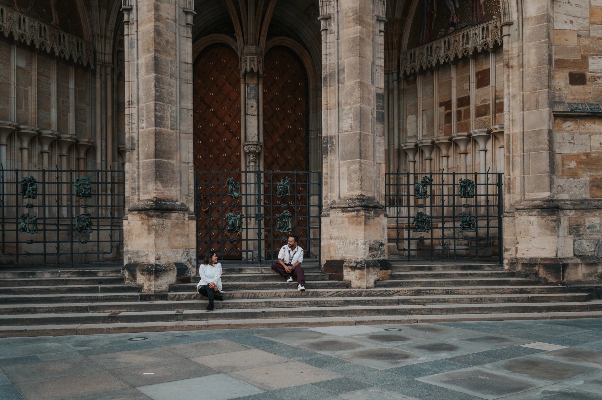 Landscape photograph of the couple sitting on the broad steps below St. Vitus Cathedral during their Prague photoshoot.