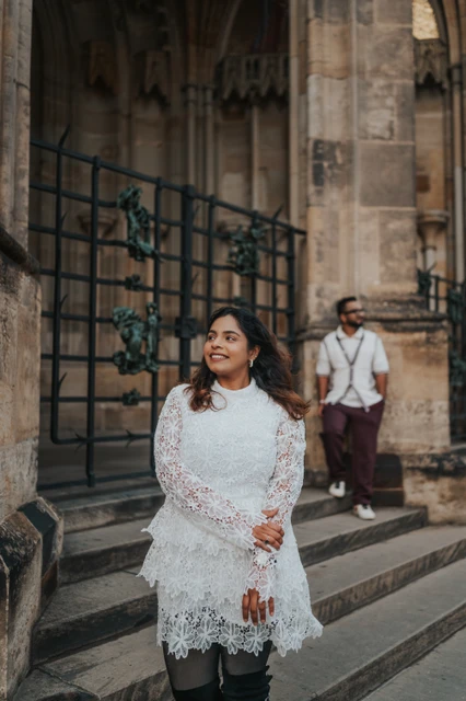 Prague photoshoot with a white lace dress by St. Vitus Cathedral Woman in a white lace dress looking to the side, with her husband on the cathedral steps behind her, during their Prague photoshoot.