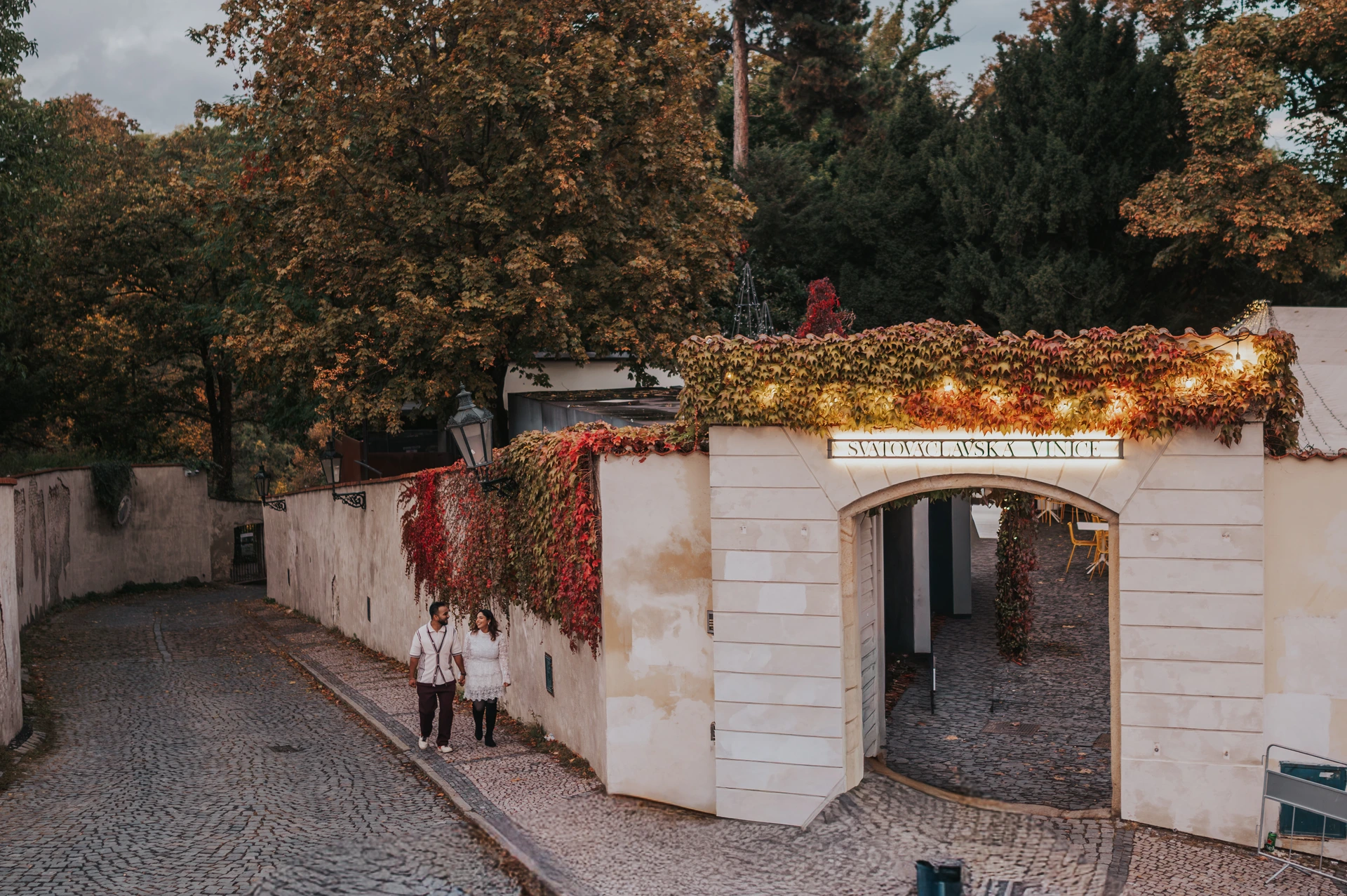 Prague couple photoshoot by the St. Wenceslas Vineyard entrance Couple walking beside autumn leaves at the St. Wenceslas Vineyard entrance during their couple photoshoot in Prague.