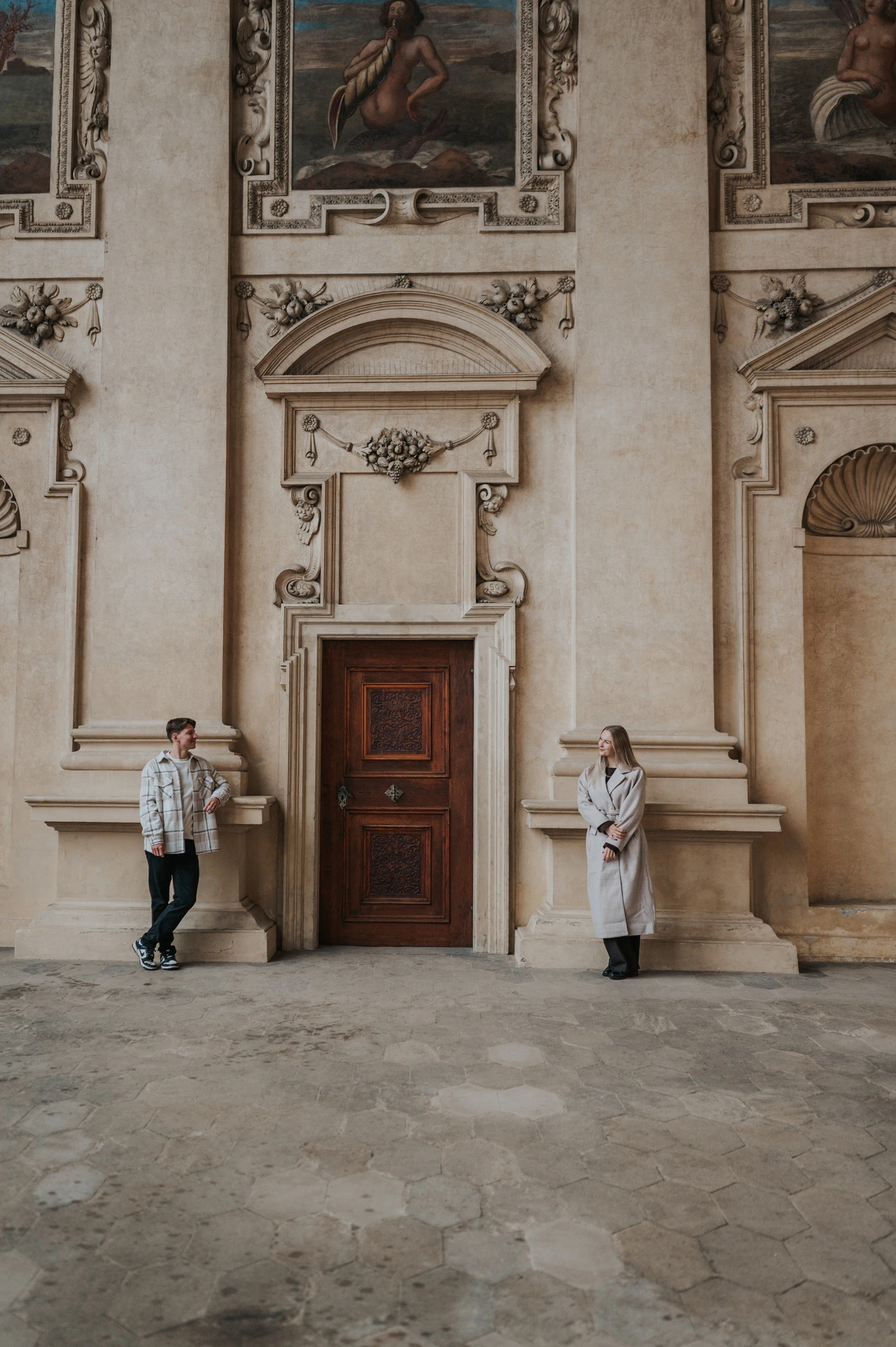 Distant portrait of the couple standing on either side of a wooden doorway beneath frescoes during their Prague couple photoshoot.