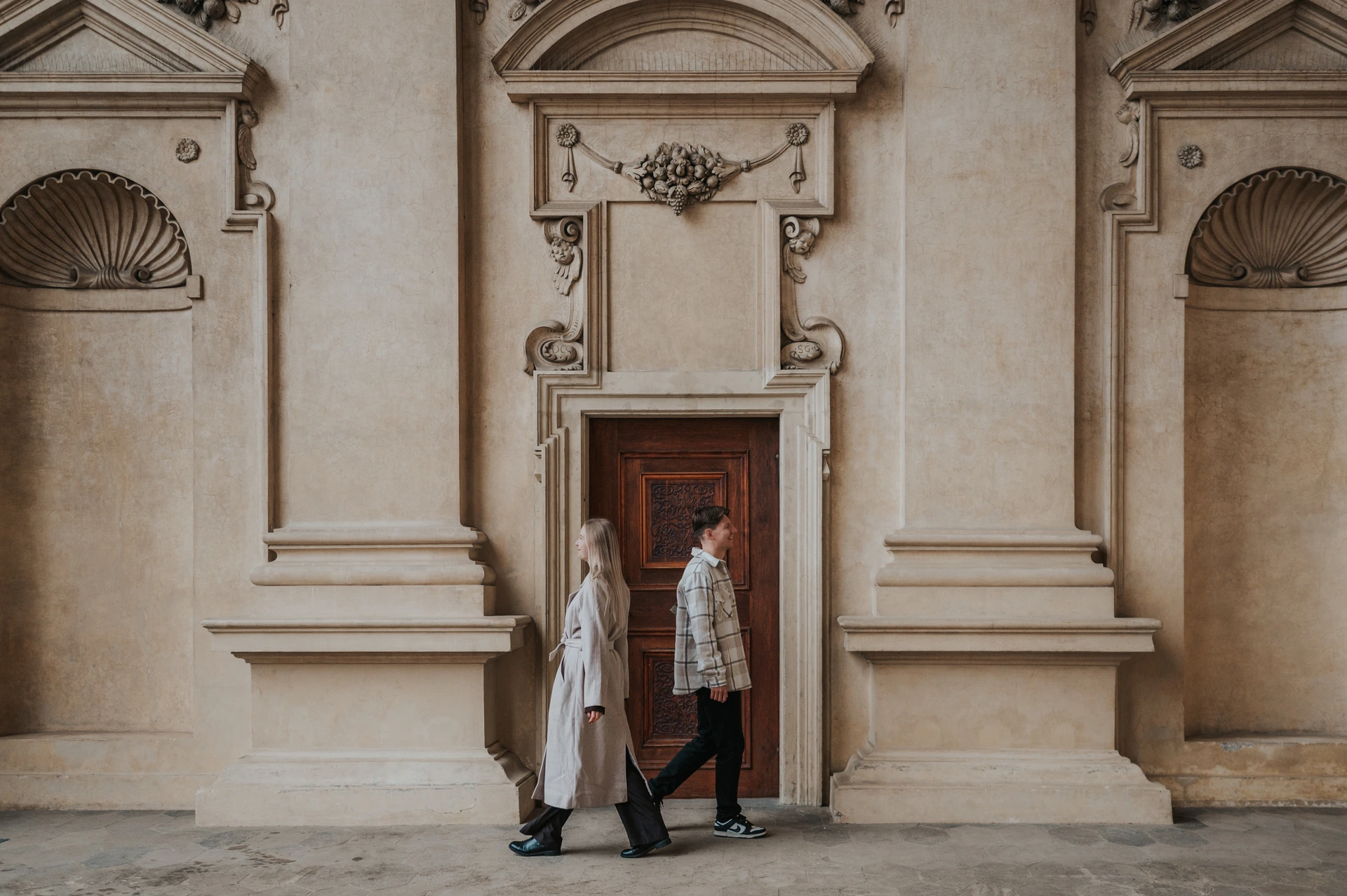 Couple walking together past a carved wooden doorway in the garden arcade during their Prague couple photoshoot.