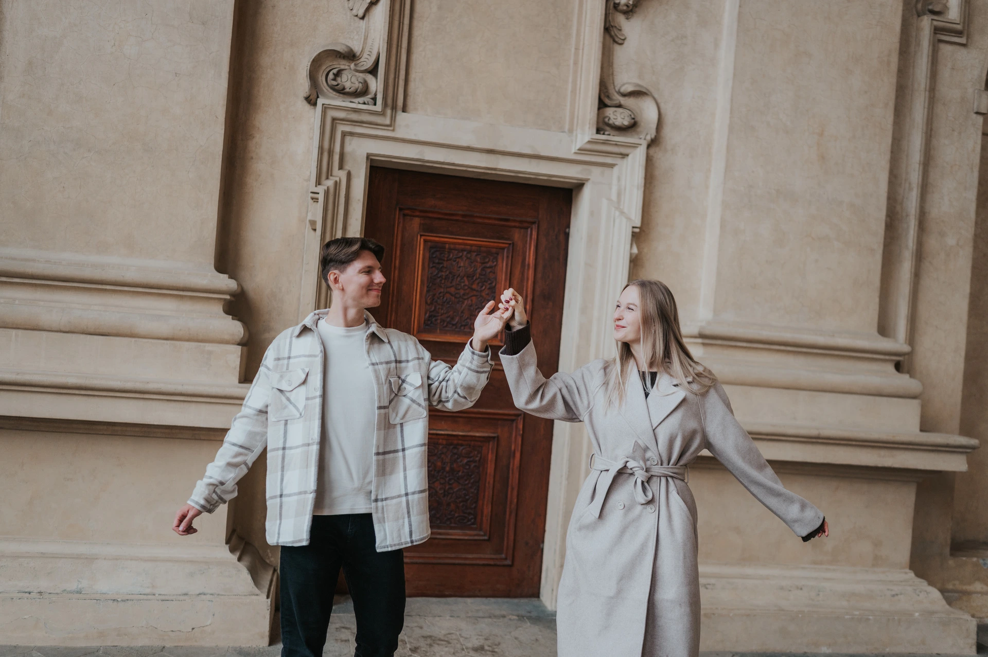 Couple stepping apart and holding hands in front of the garden arcade's wooden door during their Prague couple photoshoot.