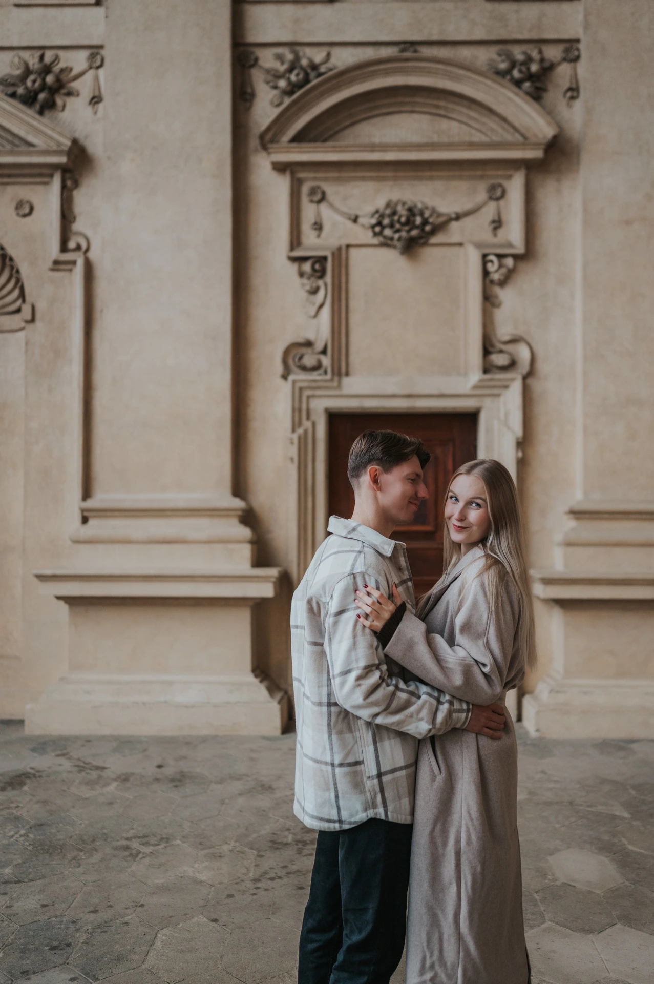 Woman smiling towards the camera as her partner embraces her in the garden arcade during their Prague couple photoshoot.