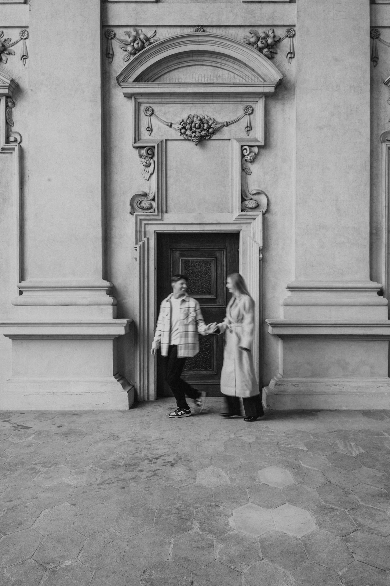 Black-and-white full-length photograph of the couple holding hands outside the arcade doorway during their Prague couple photoshoot.