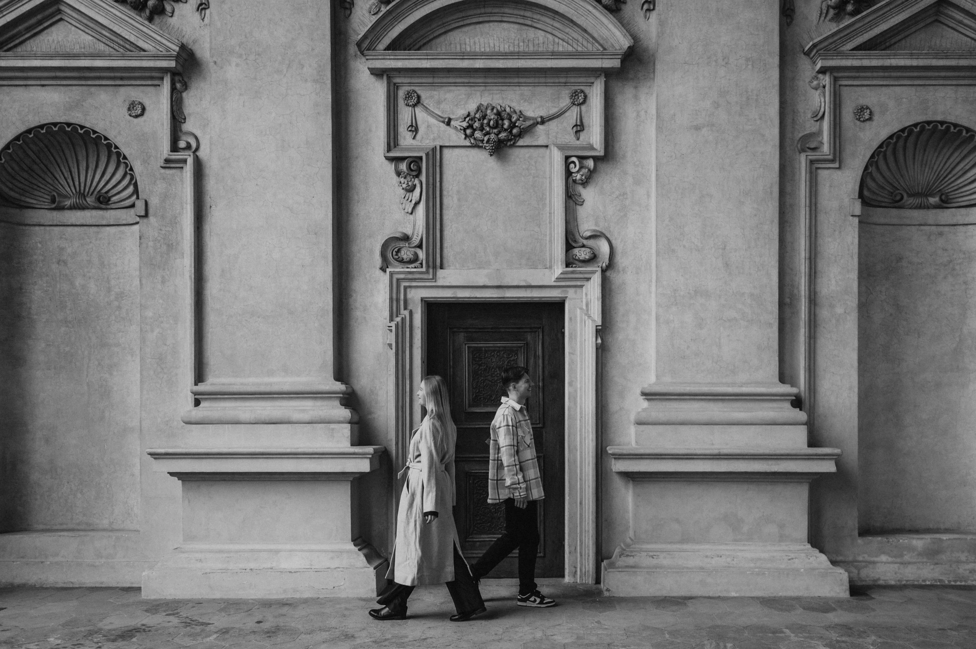 Black-and-white photograph of the couple walking past the arcade doorway during their Prague couple photoshoot.