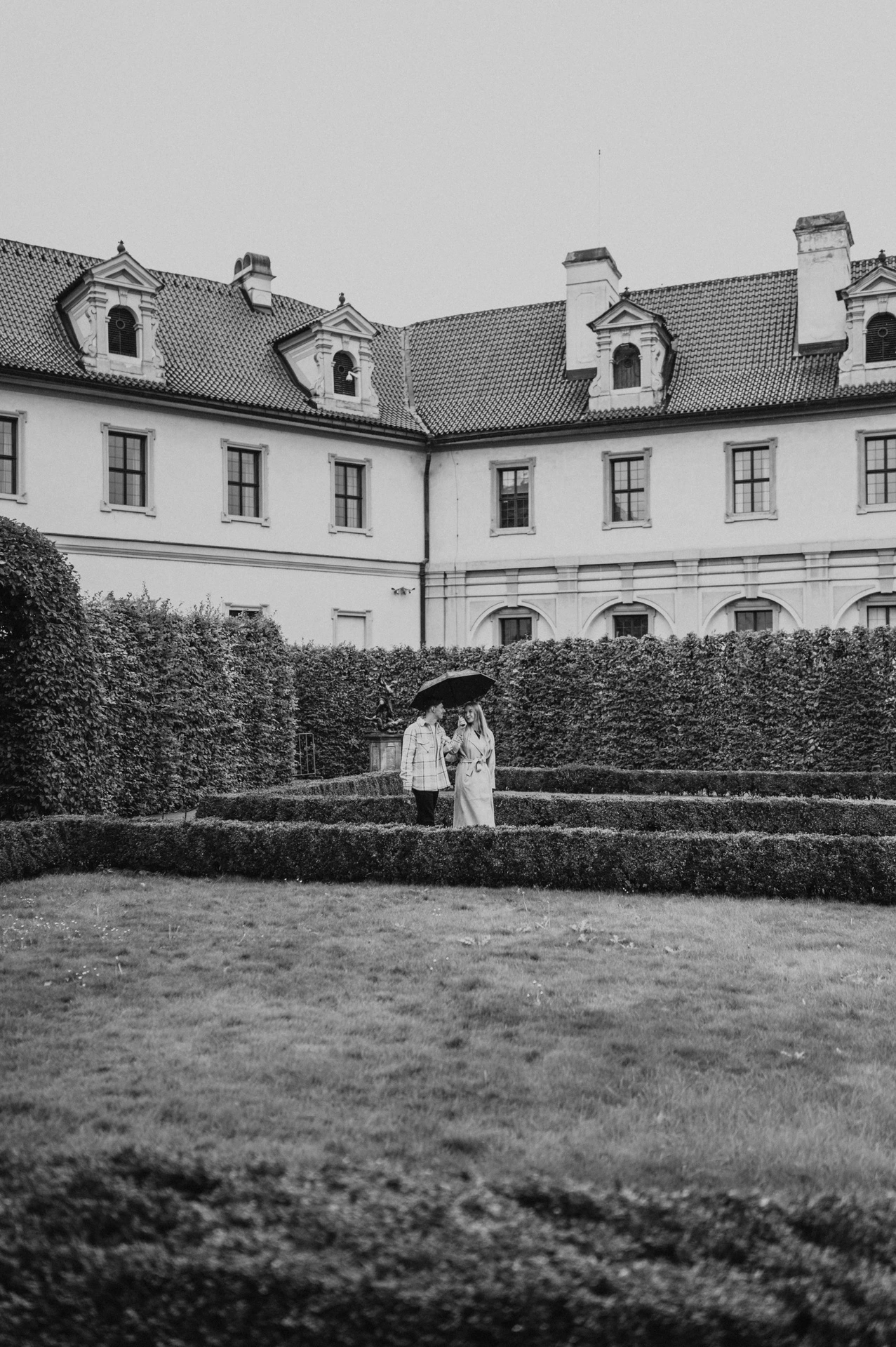 Black-and-white distant portrait of the couple beside a fountain and high garden hedges during their Prague couple photoshoot.