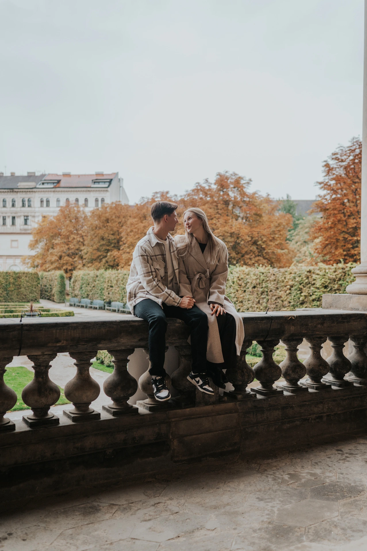 Couple kissing while seated on a garden balustrade with autumn trees behind them during their Prague couple photoshoot.