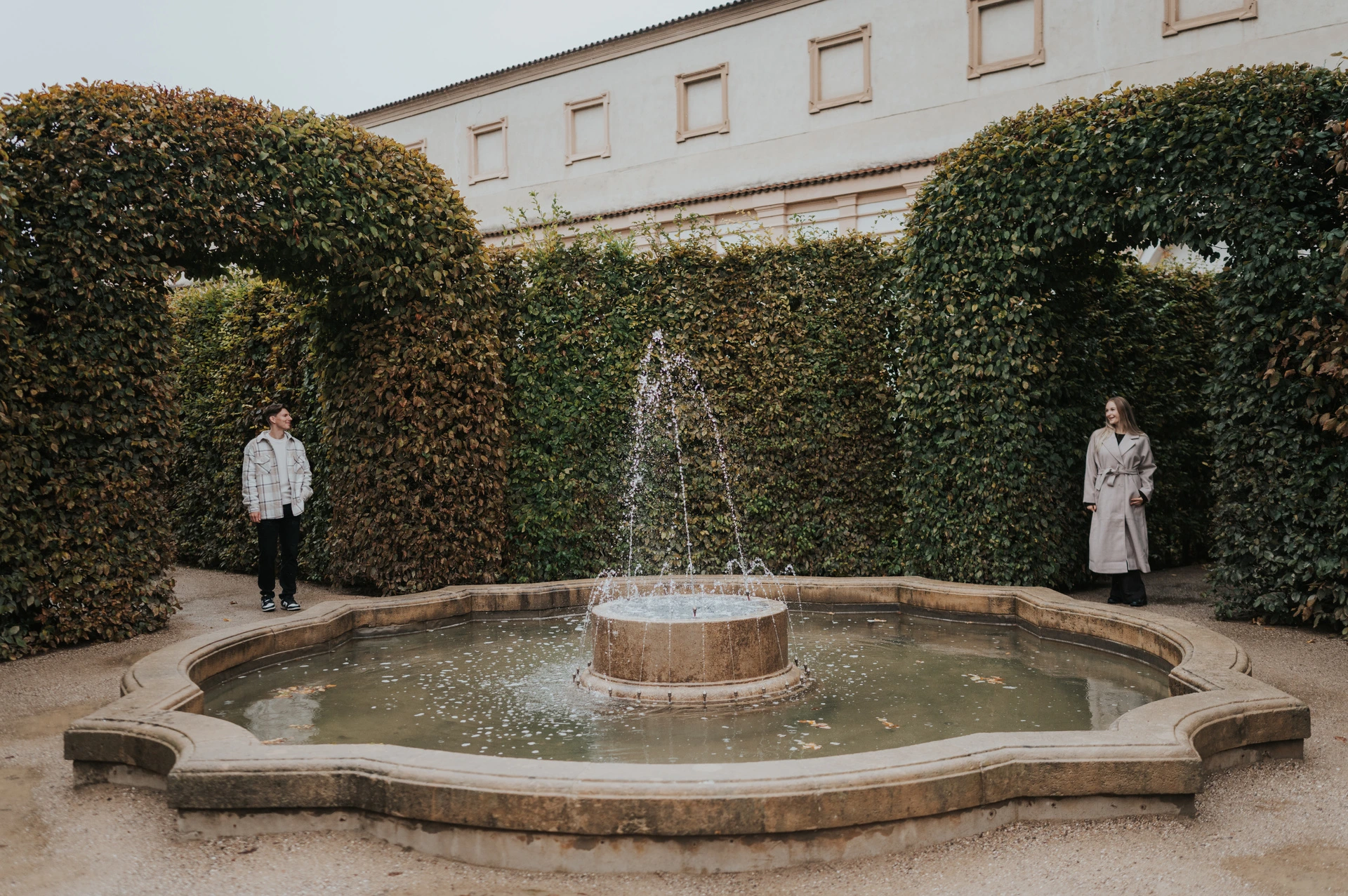Couple standing on either side of a small fountain surrounded by tall green hedges during their Prague couple photoshoot.