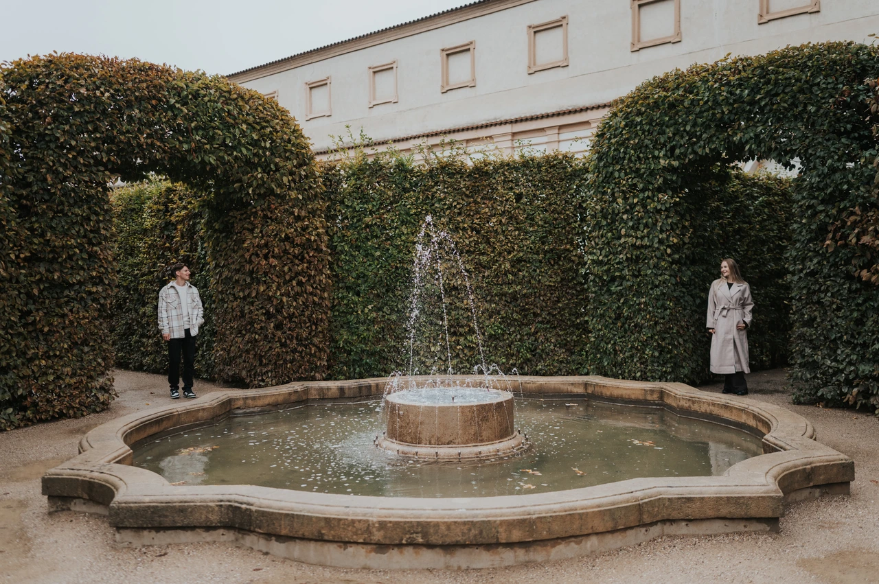 Prague couple photoshoot: two sides of a fountain in Wallenstein Garden Couple standing on either side of a small fountain surrounded by tall green hedges during their Prague couple photoshoot.