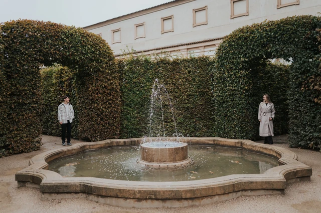 Couple standing on either side of a small fountain surrounded by tall green hedges during their Prague couple photoshoot.