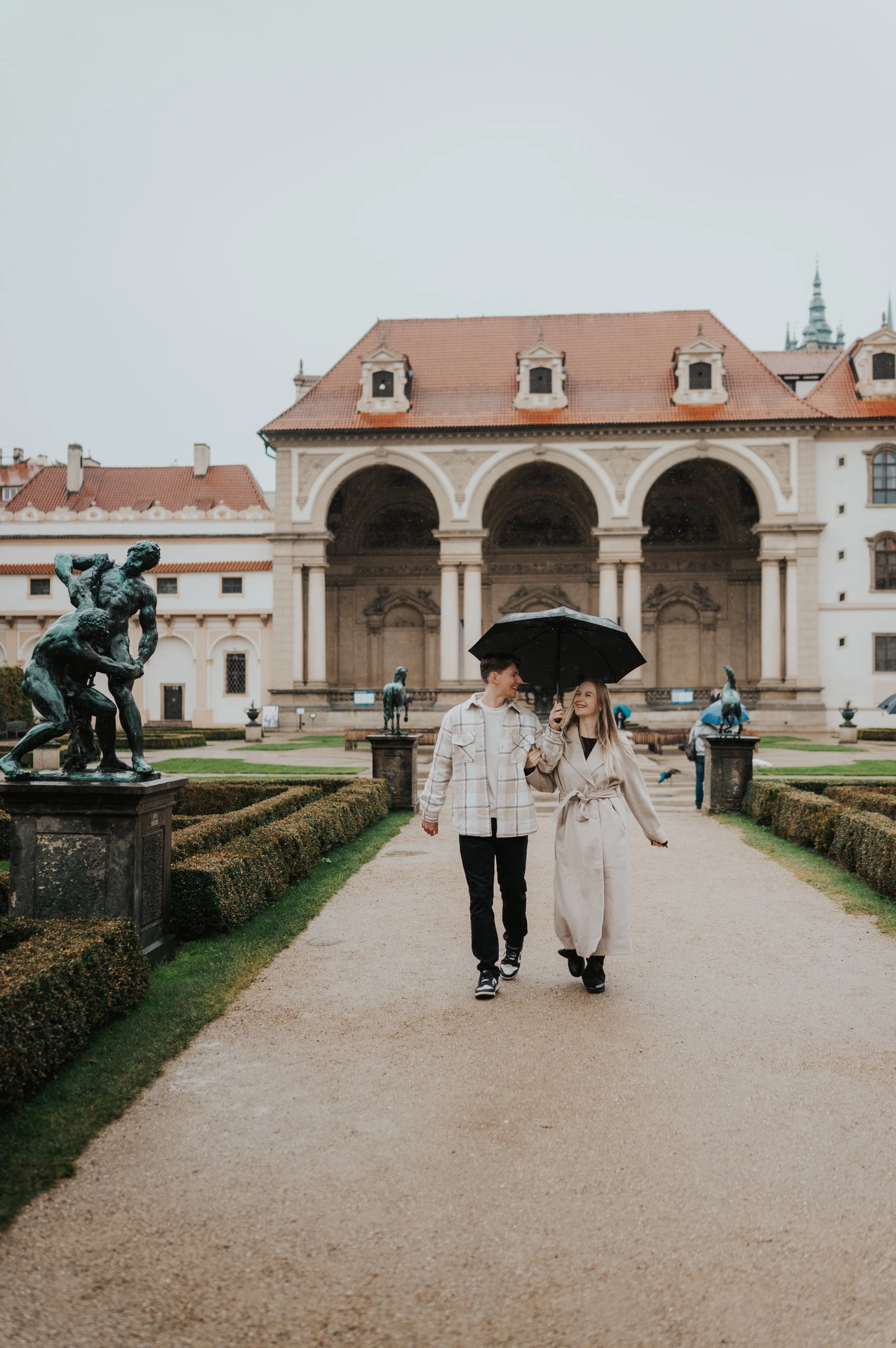 Couple walking hand in hand down the formal garden path beneath a shared umbrella during their Prague couple photoshoot.