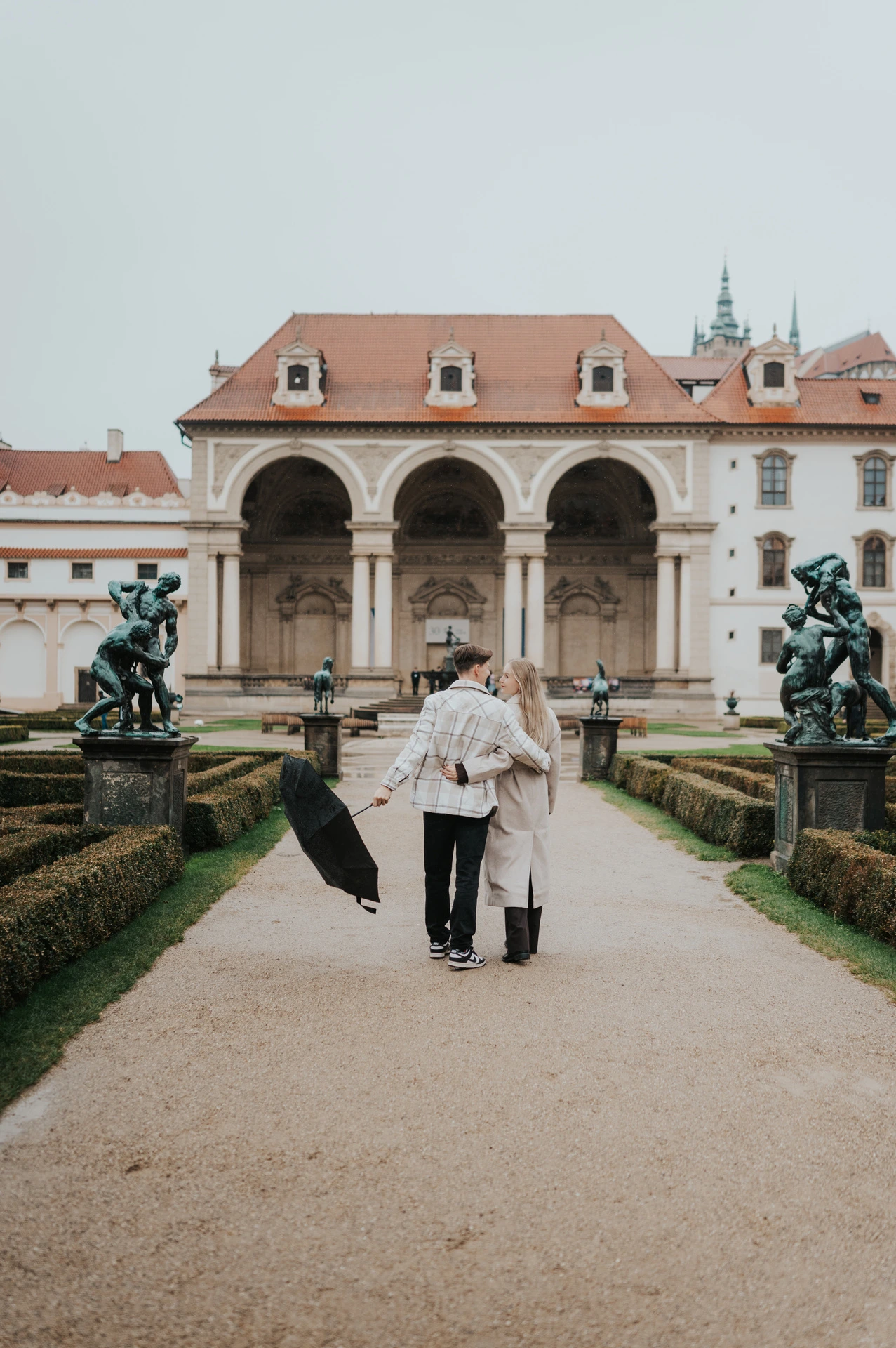Couple walking together on the garden path with their umbrella held to one side during their Prague couple photoshoot.