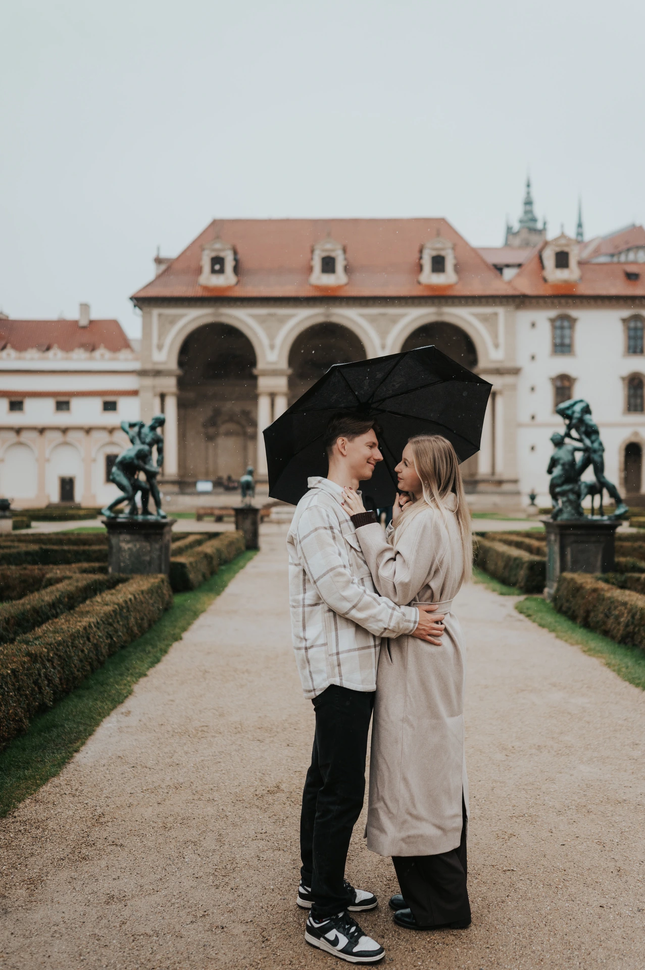 Couple embracing beneath a black umbrella with the garden loggia in the background during their Prague couple photoshoot.