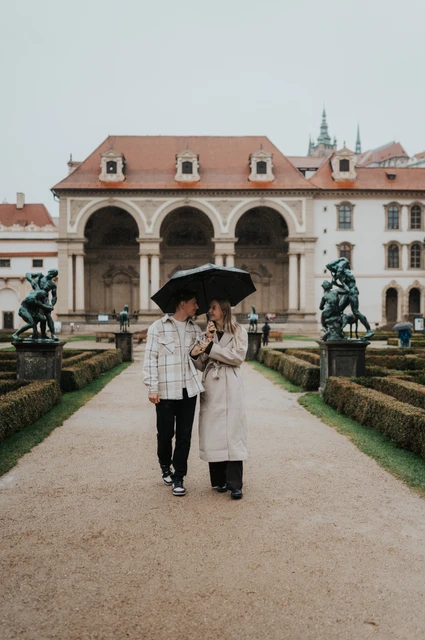 Prague couple photoshoot: under an umbrella in Wallenstein Garden Couple sharing a black umbrella on a garden path with the three-arched loggia behind them during their Prague couple photoshoot.