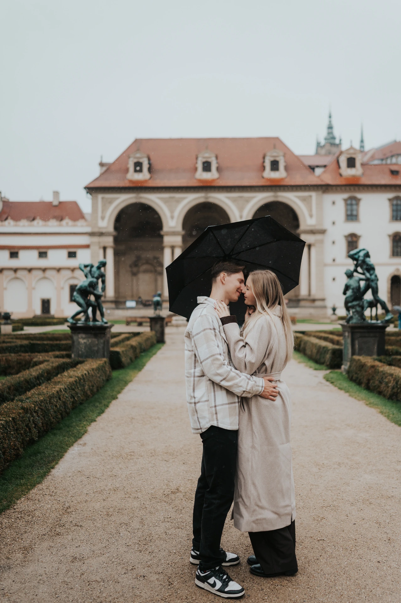 Couple kissing under their black umbrella on a formal garden path during their Prague couple photoshoot.