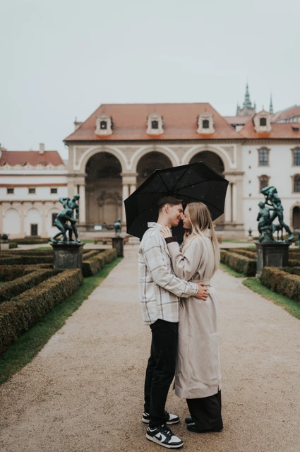 Prague couple photoshoot: a rainy garden kiss beneath the umbrella Couple kissing under their black umbrella on a formal garden path during their Prague couple photoshoot.