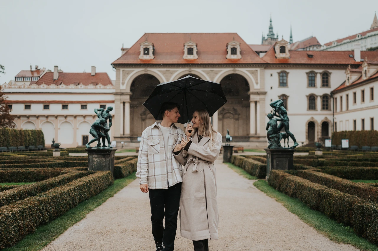 Prague couple photoshoot: a shared umbrella beside the formal hedges Wide view of the couple sharing an umbrella between formal hedges in front of the garden loggia during their Prague couple photoshoot.