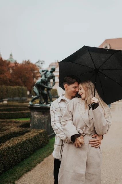 Prague couple photoshoot: a cheek kiss under the garden umbrella Man kissing his partner's cheek as she smiles beneath their black umbrella during their Prague couple photoshoot.