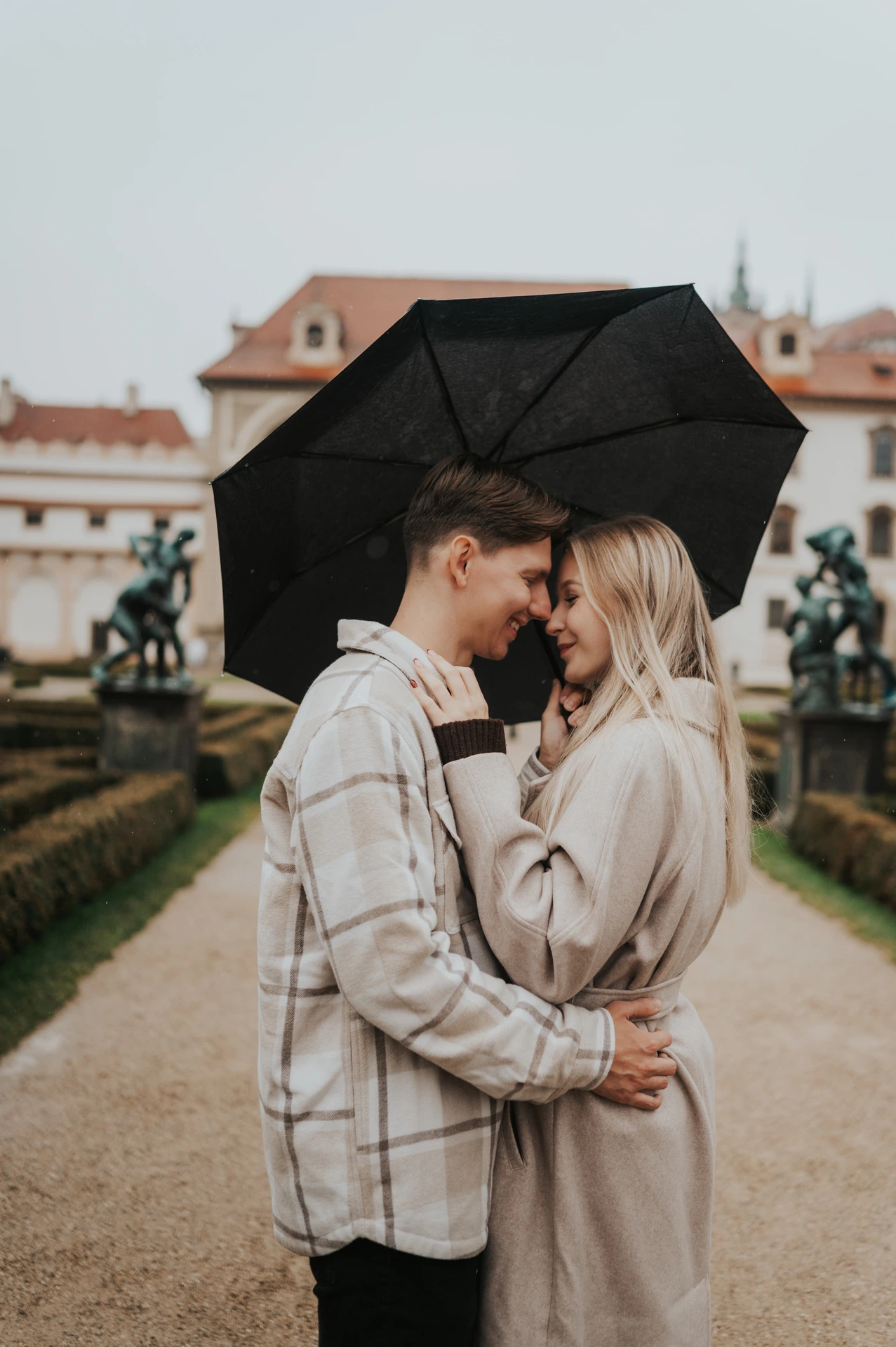 Couple touching foreheads under a black umbrella beside clipped garden hedges during their Prague couple photoshoot.
