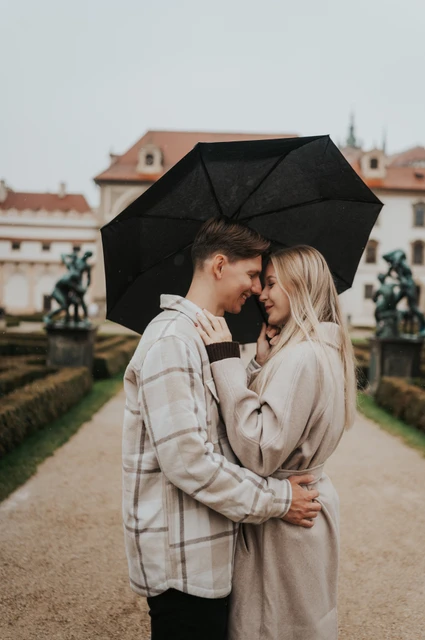 Prague couple photoshoot: foreheads together under the umbrella Couple touching foreheads under a black umbrella beside clipped garden hedges during their Prague couple photoshoot.